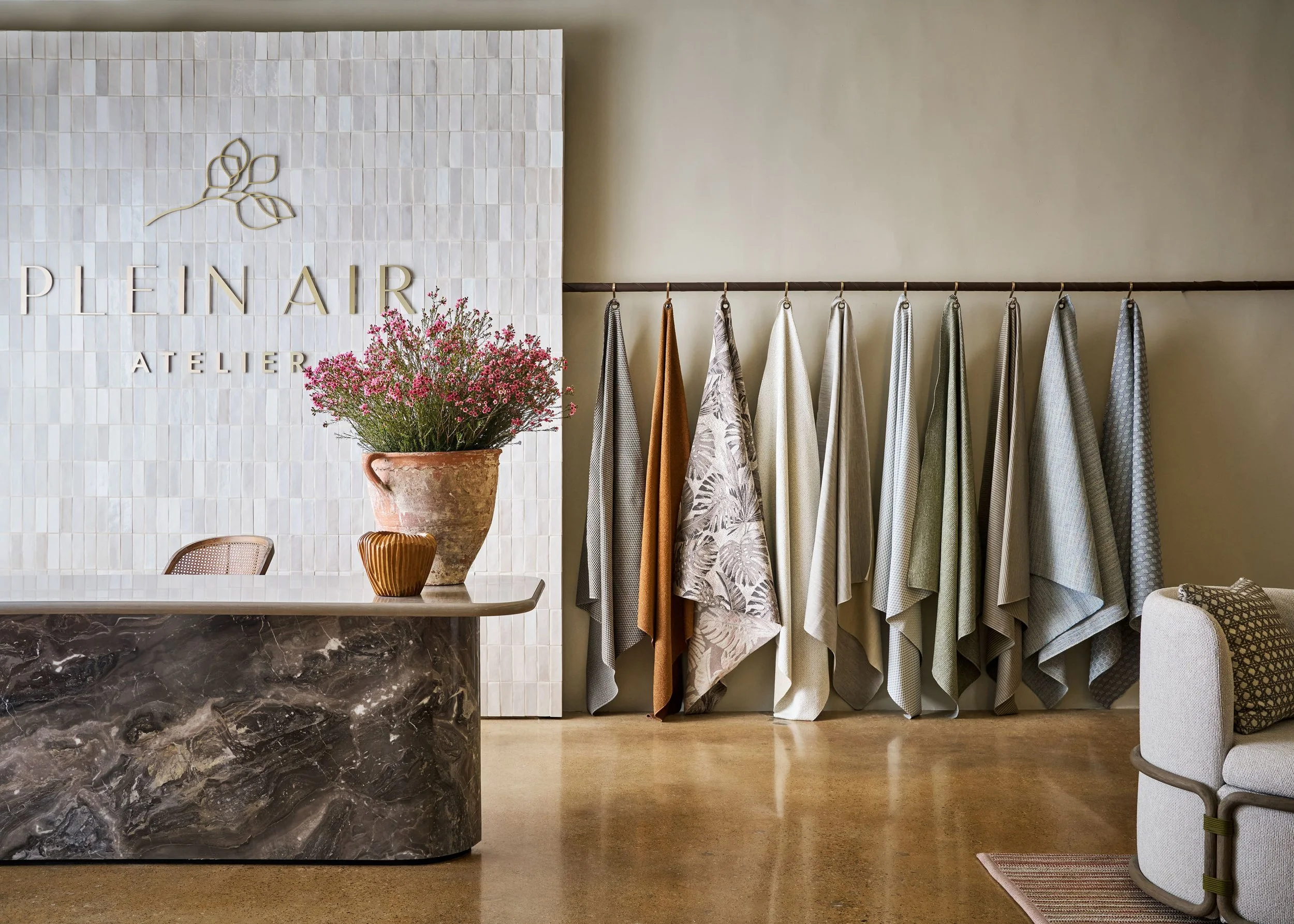 Interior of a fabric showroom with a wall displaying textured fabrics hanging on a rod, a large pot with pink flowers on a marble counter, and a sign reading 'Plein Air Atelier' on a tiled wall.