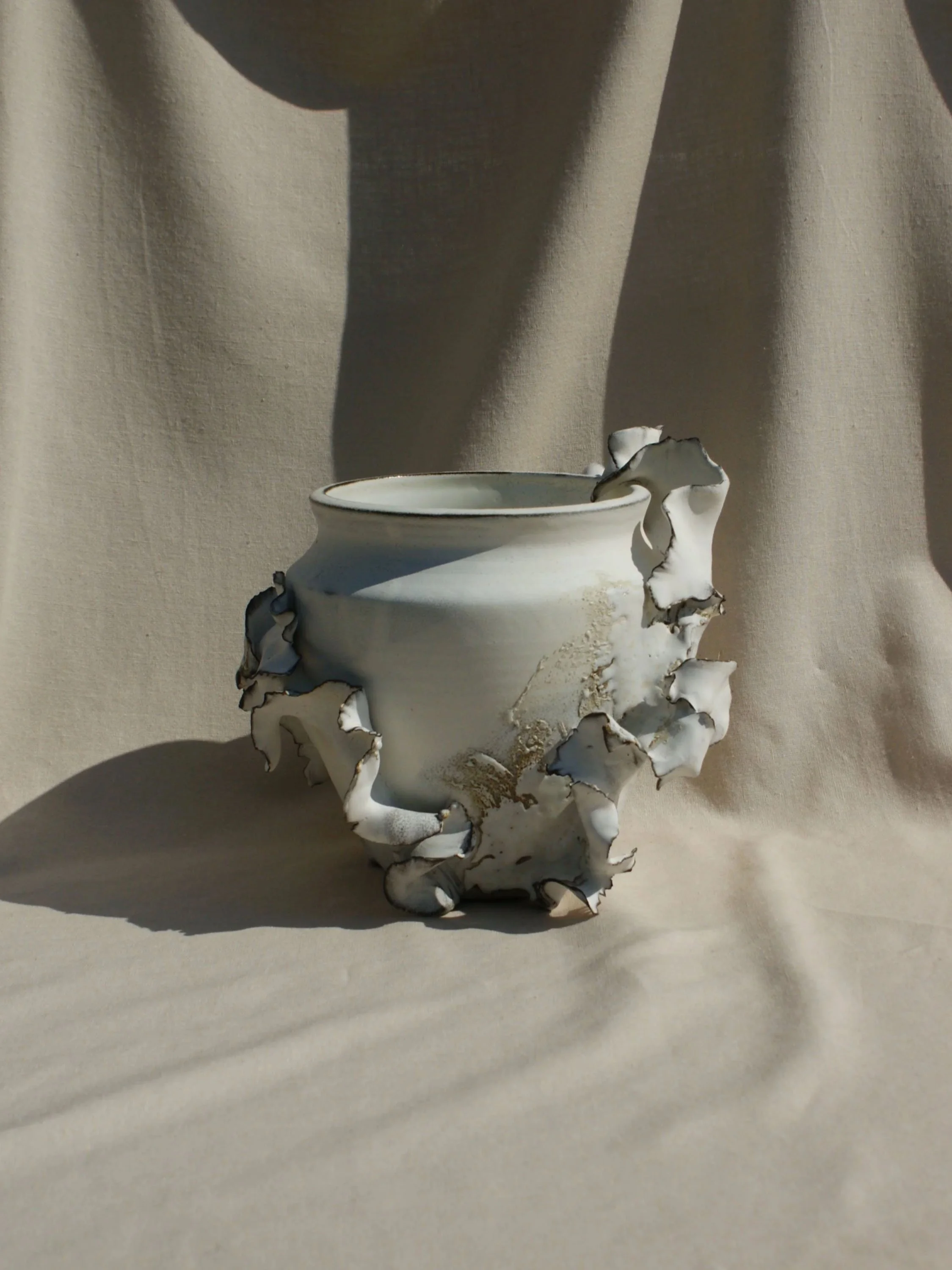 A white ceramic vessel with irregular ruffled edges and gold accents, placed against a beige fabric background.