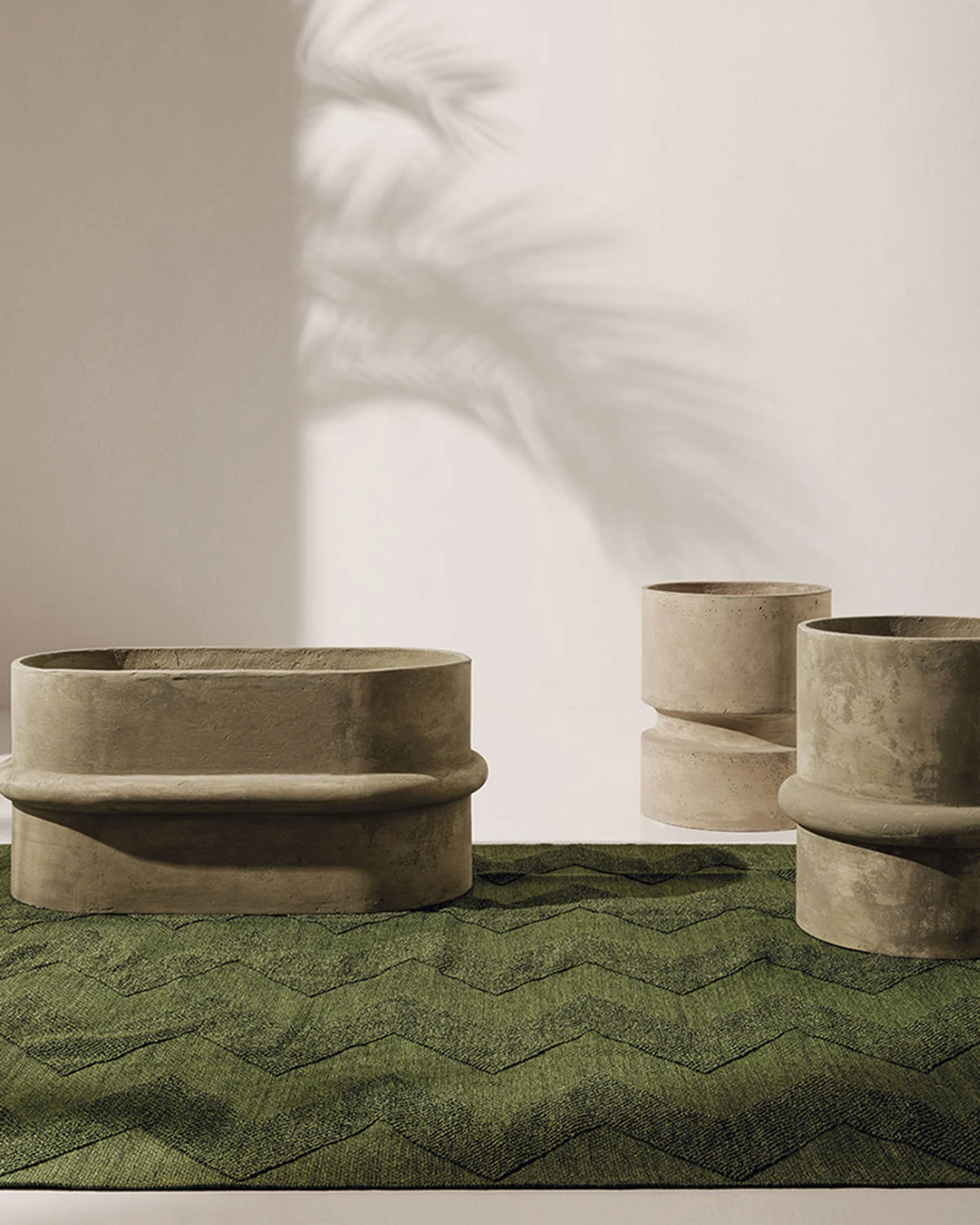 Three beige concrete planters of varying sizes arranged on green textured surface, with shadow of plant cast on plain light-colored wall in background.