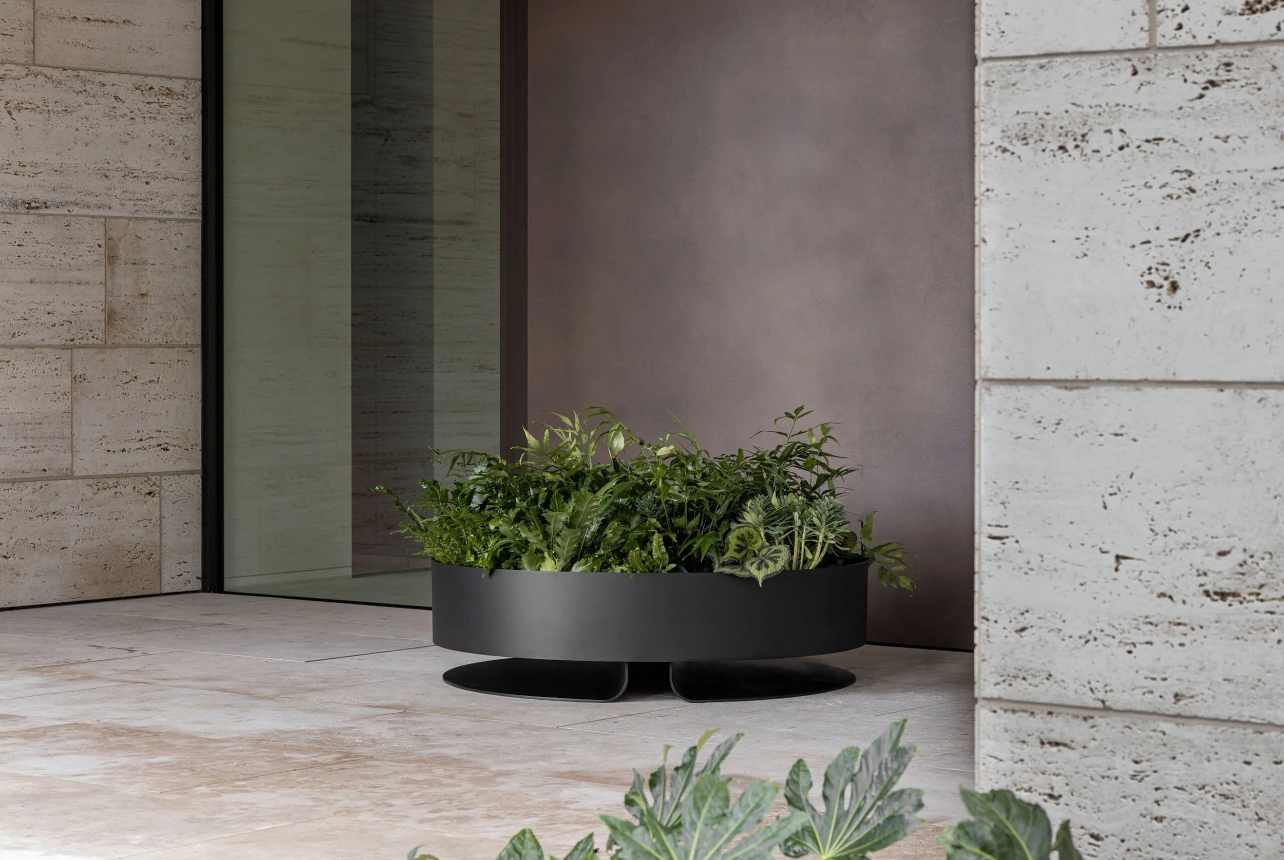 A black planter with green leafy plants outside a modern building with stone and concrete walls.