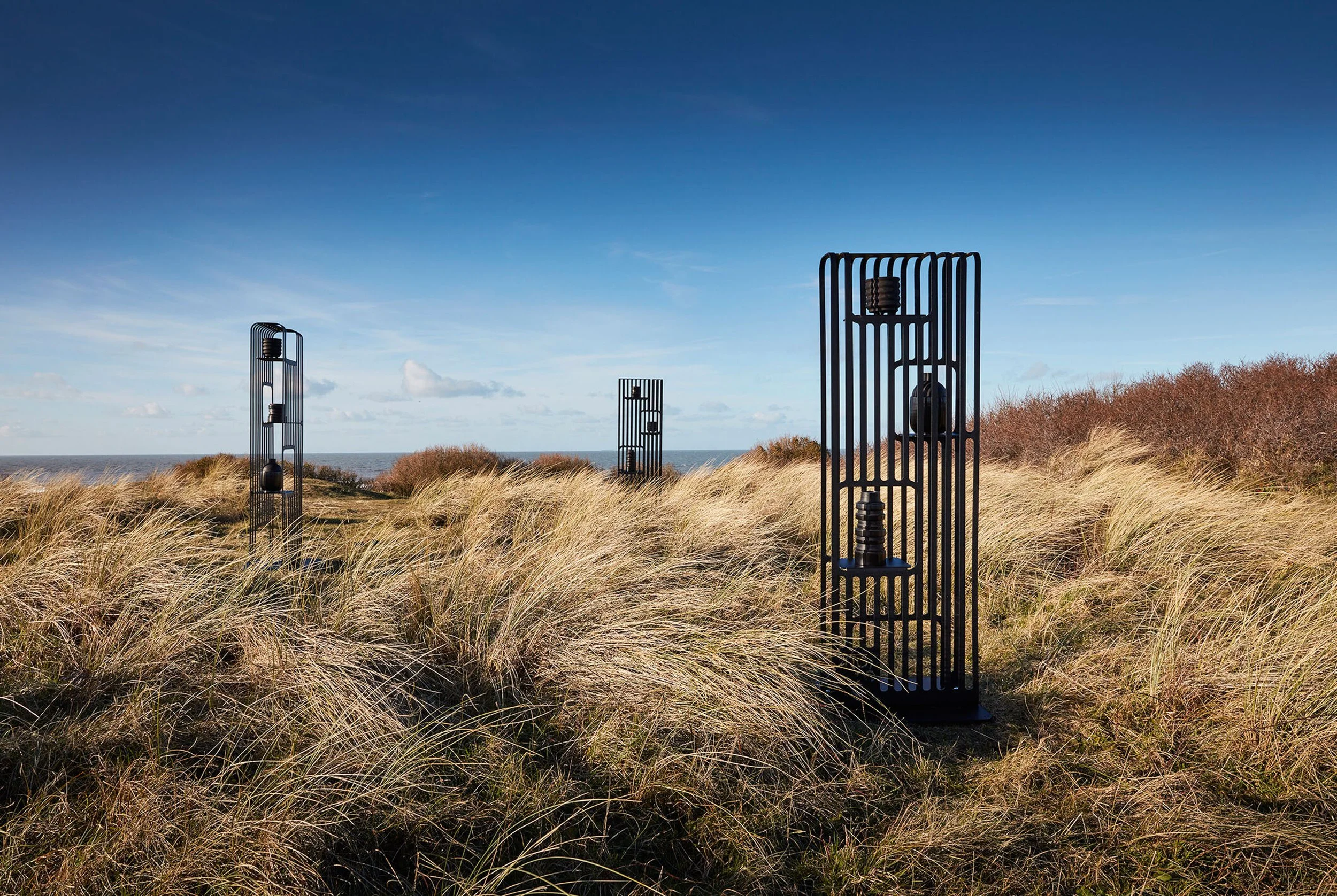 Beach landscape with tall grass and a clear blue sky, featuring three black metal sculptures resembling elongated Mailboxes.