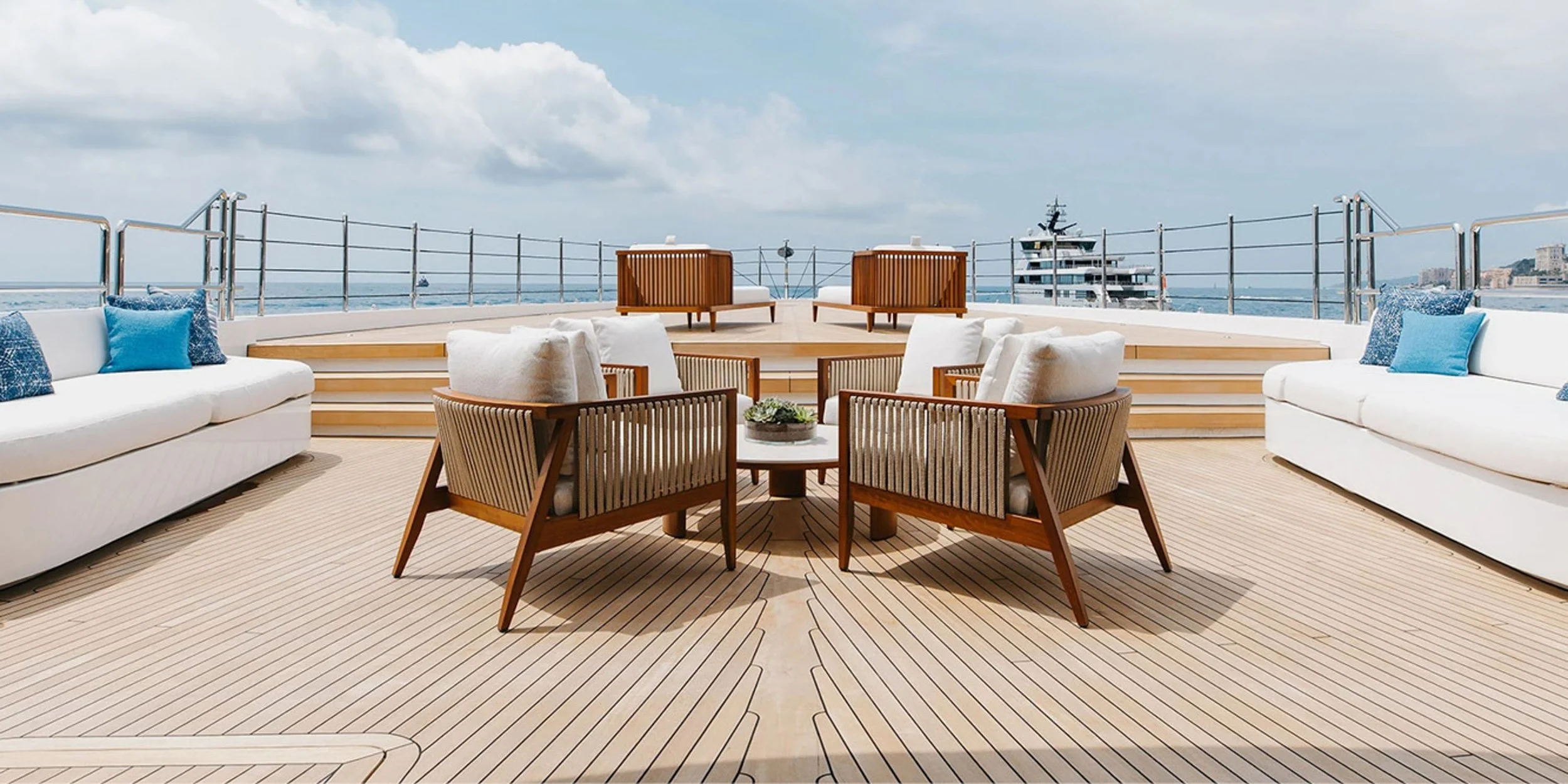 A luxurious outdoor lounge area on a yacht with white sofas, wooden chairs, and a view of the sea and a yacht in the background.