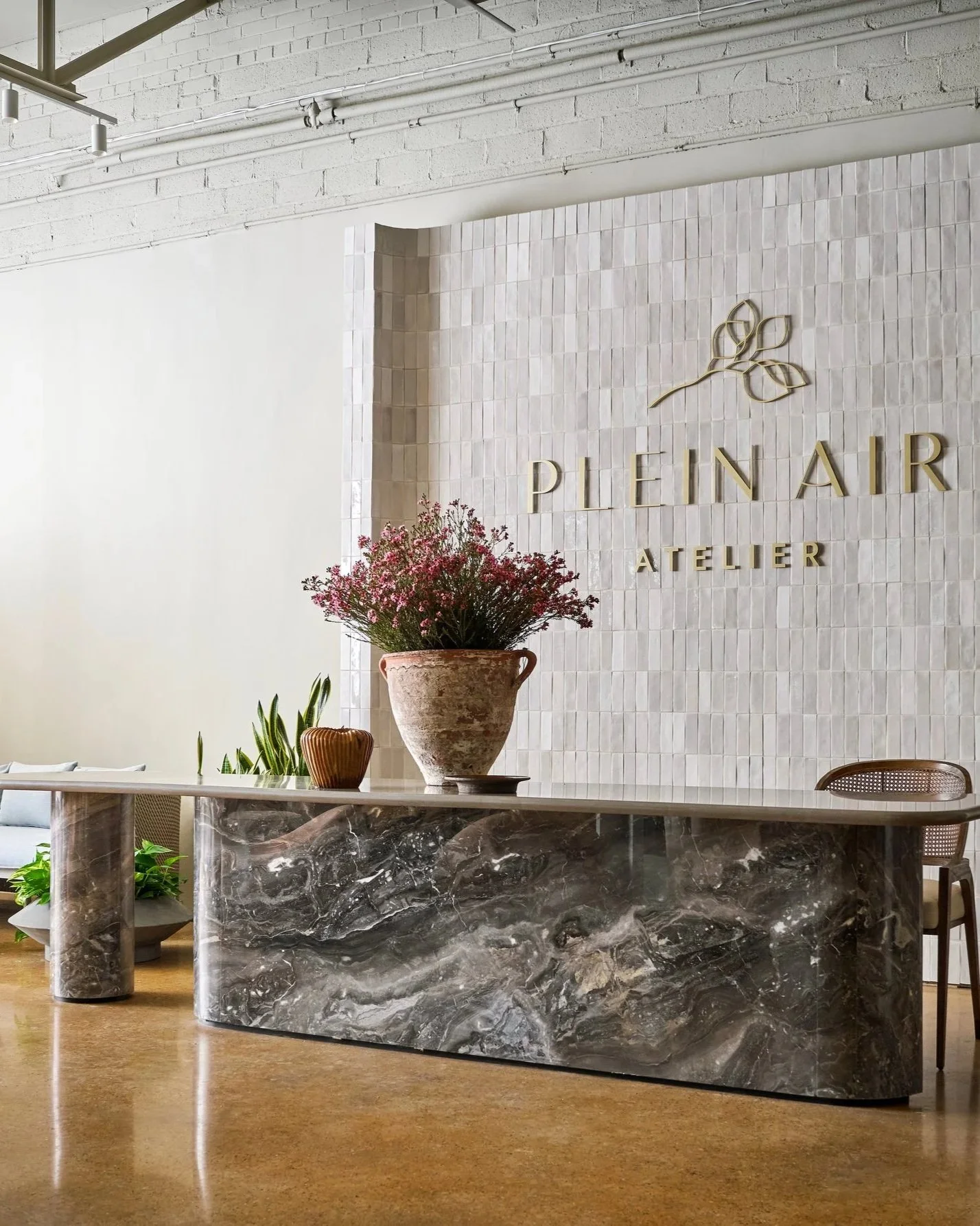 Modern reception area with a marble desk, large potted plant, and decorative objects. White textured wall features the logo 'Plein Air Atelier' with a leaf design.