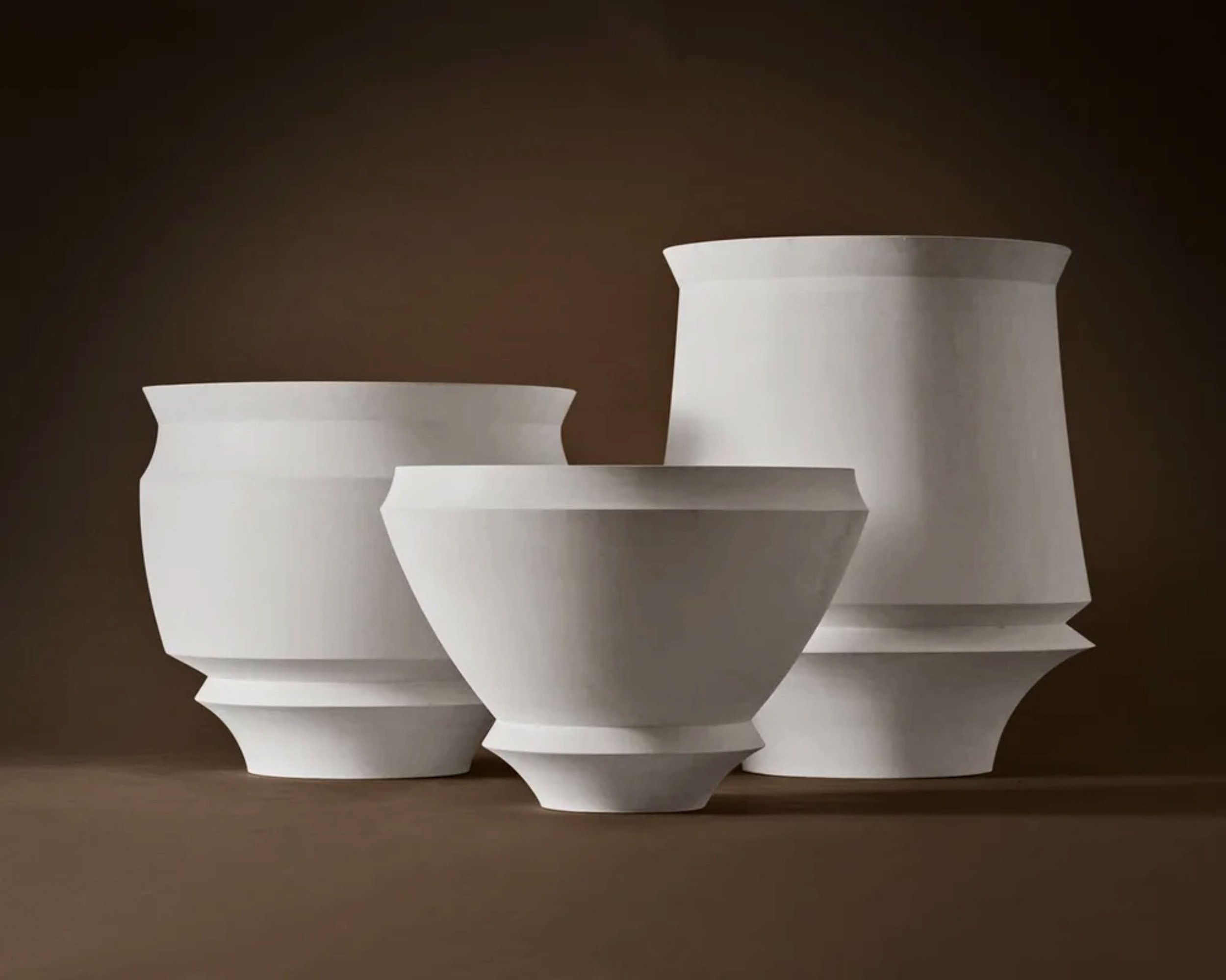 Three white, tiered vases with curved, flared openings lined up on a wooden surface against a dark background.