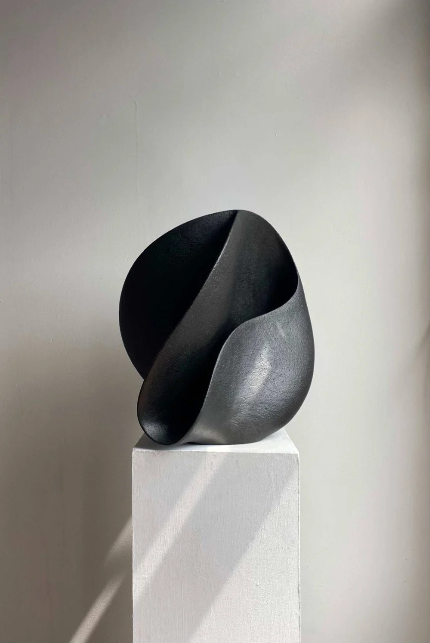 Abstract black and gray sculpture placed on a white pedestal against a plain wall.