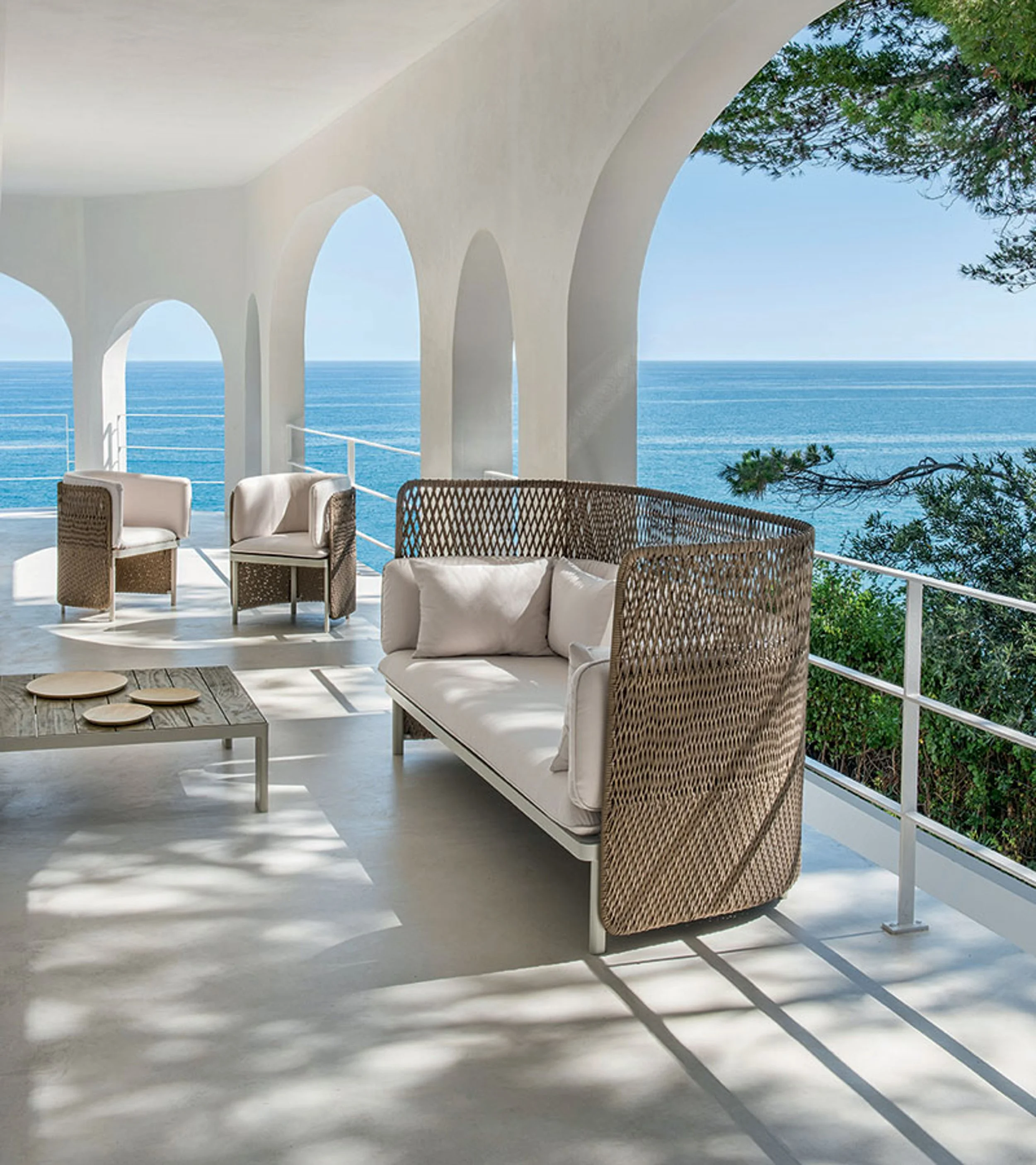 Beachside balcony with white walls and arched openings, wicker and cushioned chairs, a rustic table, and a view of the blue ocean and sky with a green tree branch in the foreground.