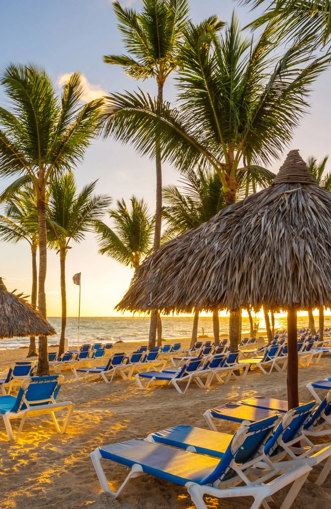 A beach with blue loungers and cabanas and palm trees overlooking a Caribbean sunset.