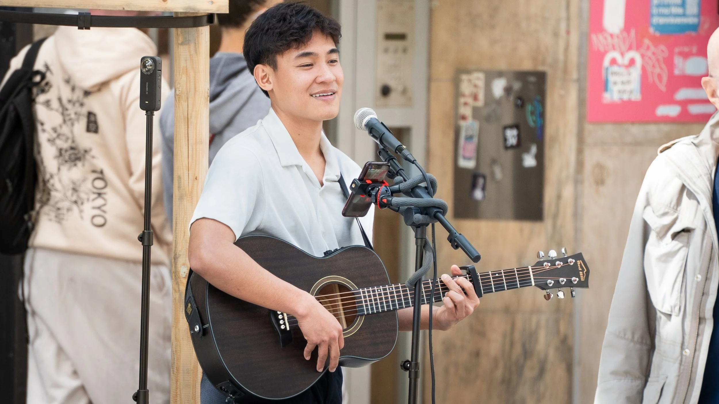 Jeune homme jouant de la guitare et chantant dans un lieu public, avec un microphone et plusieurs personnes autour.