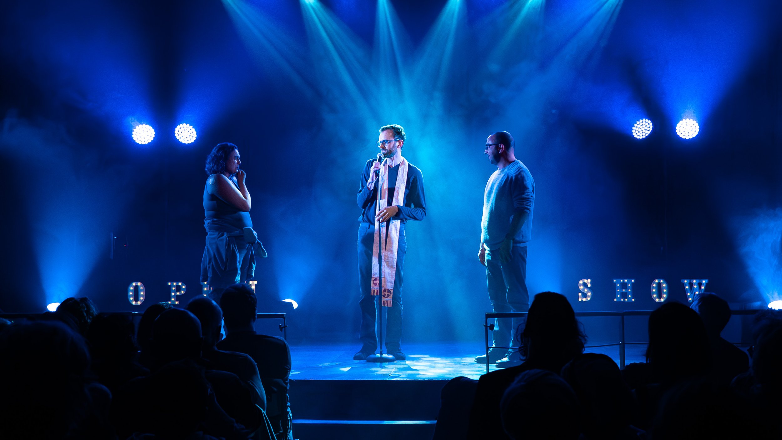 Trois personnes se tiennent sur une scène sous des lumières bleues, l'une d'elles parle dans un microphone, lors d'un spectacle ou d'une présentation, avec l'audience visible en silhouette devant la scène.