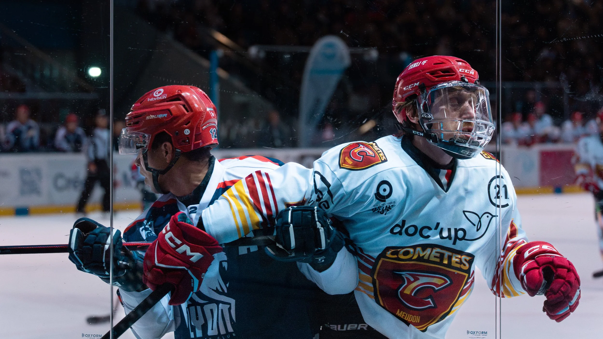 Deux joueurs de hockey en compétition, un portant un casque rouge et un maillot blanc avec des logos, l'autre portant un casque rouge et un maillot blanc avec des logos, se battent sur la glace lors d'un match.