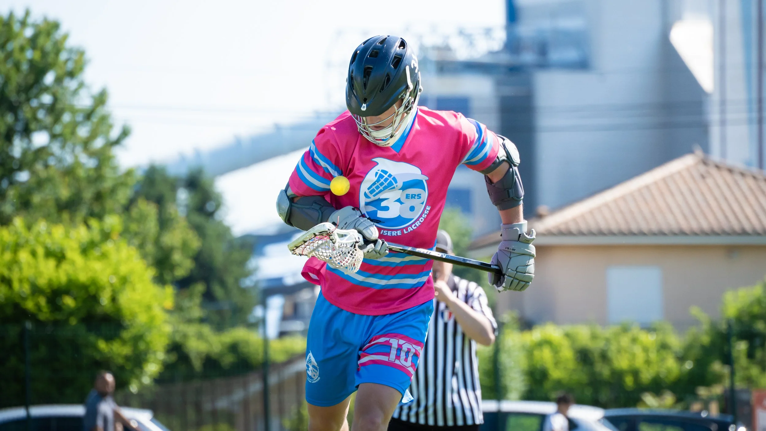 Un joueur de crosse en tenue rose et bleue, portant un casque noir et un protège-bras, tient une crosse de hockey en regardant une balle jaune pendant un match en extérieur.
