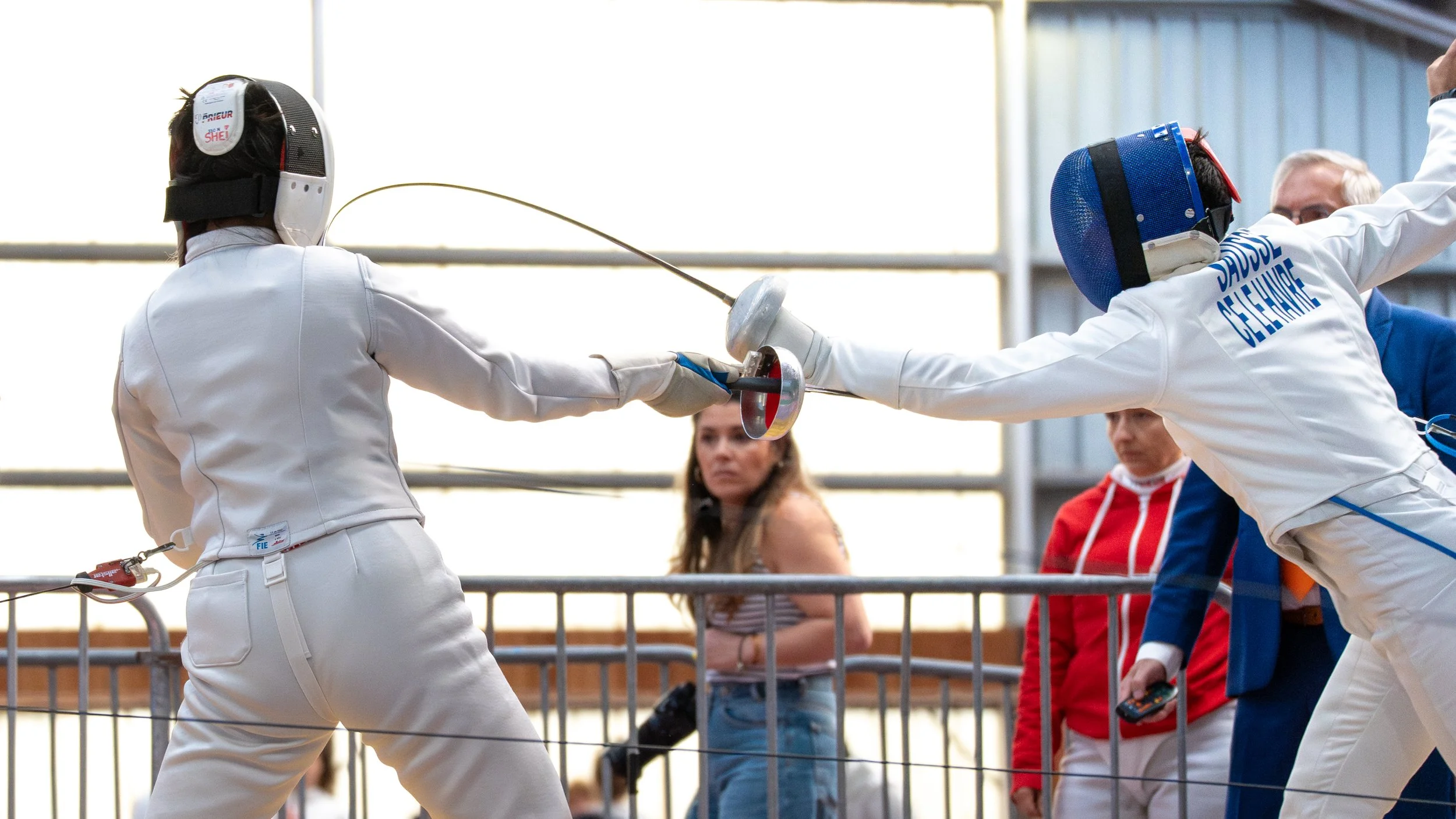 Deux escrimeuses en combat, portant des sallettes et des blouses blanches, s'affrontent à l'épée, avec une personne en arrière-plan et un public en spectateur.