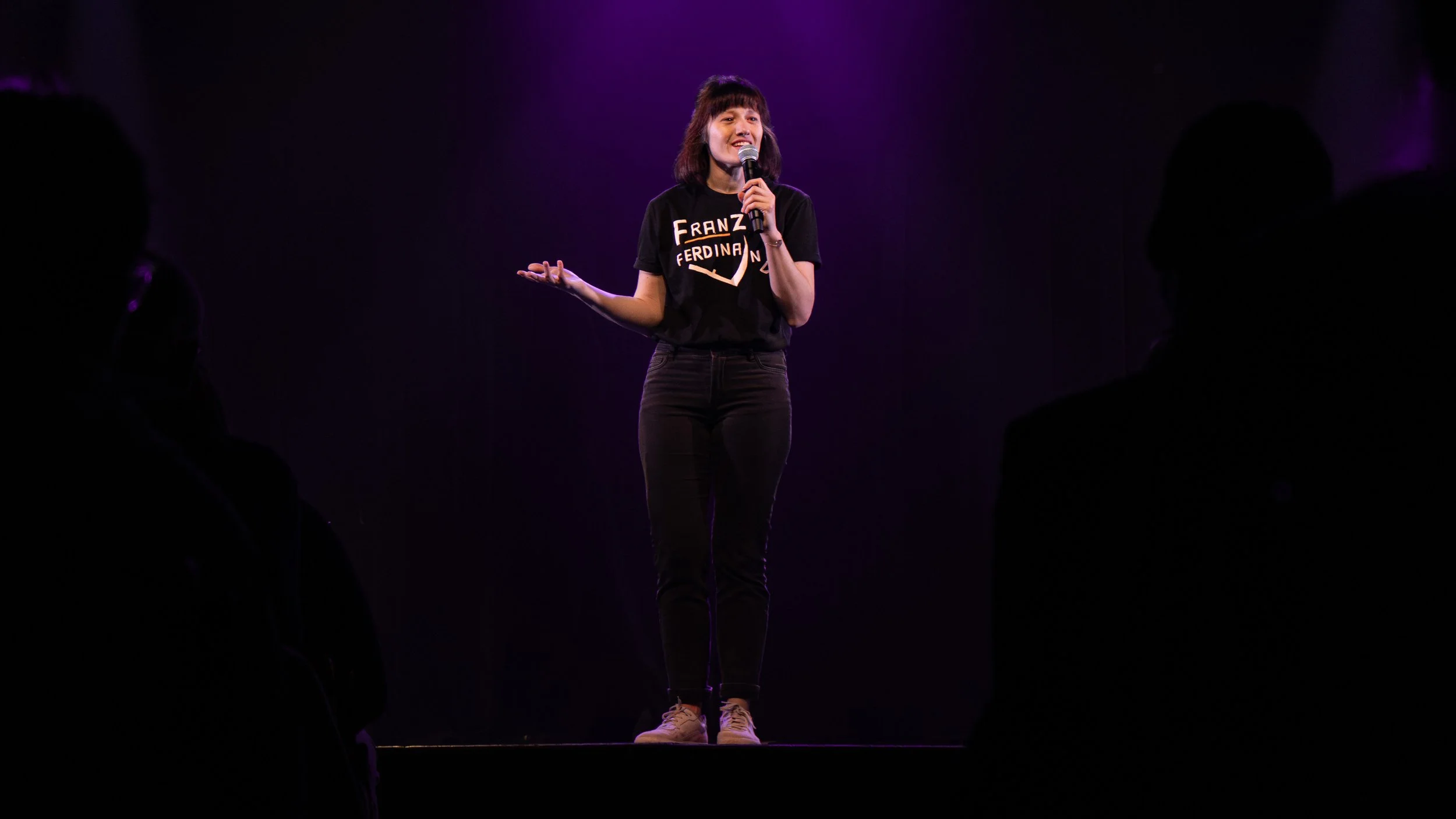 Une femme parle au micro sur scène, portant un t-shirt noir avec des mots en français, dans une ambiance sombre avec un éclairage violet, vue par un public silhouette