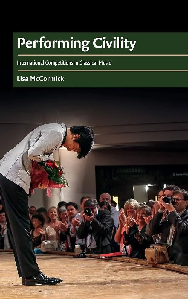 A young man bows on stage, holding a bouquet of flowers, as an audience claps and takes photos, celebrating a performance or achievement.