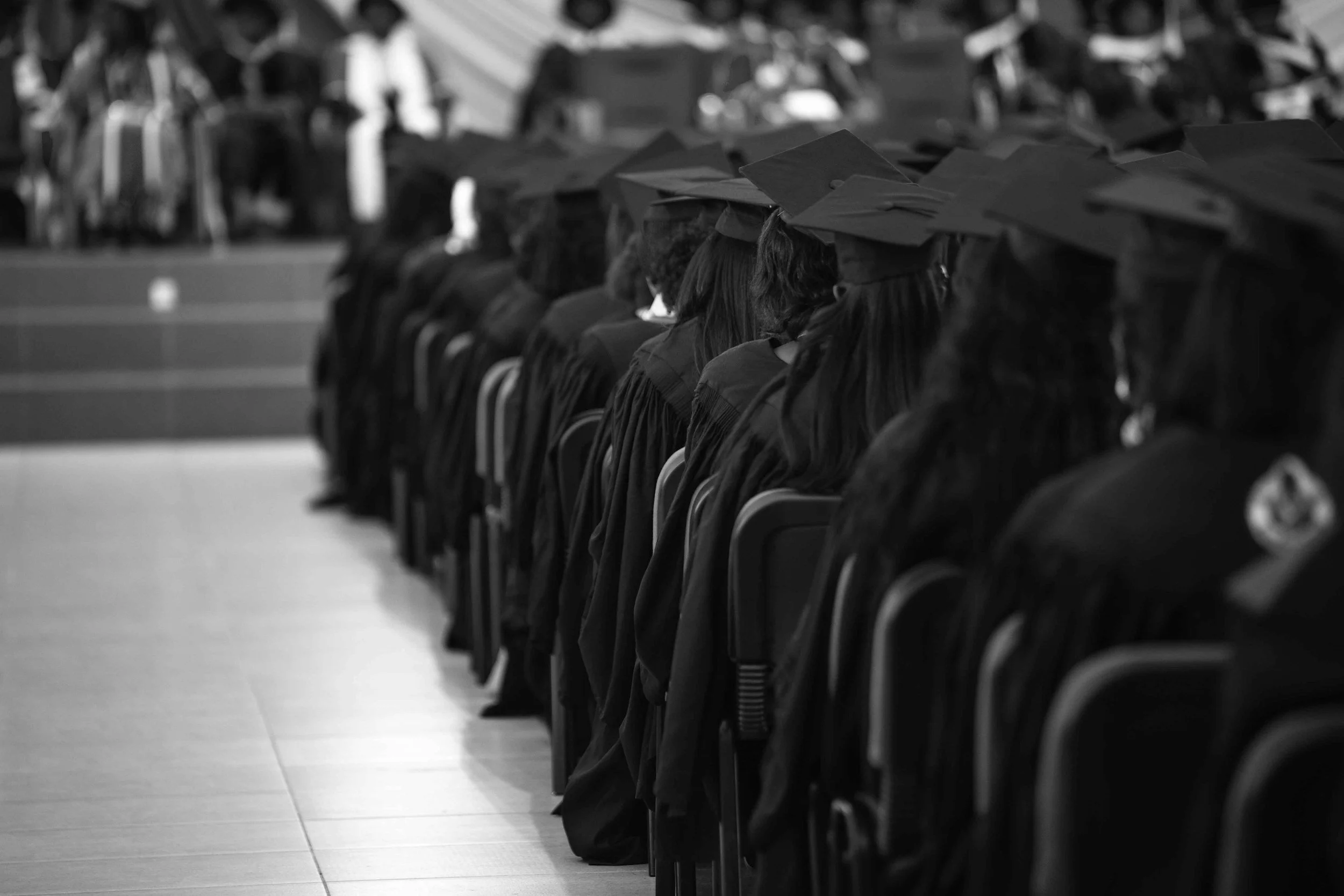 Black and white photo of multiple graduates seated in rows at a graduation ceremony wearing caps and gowns.