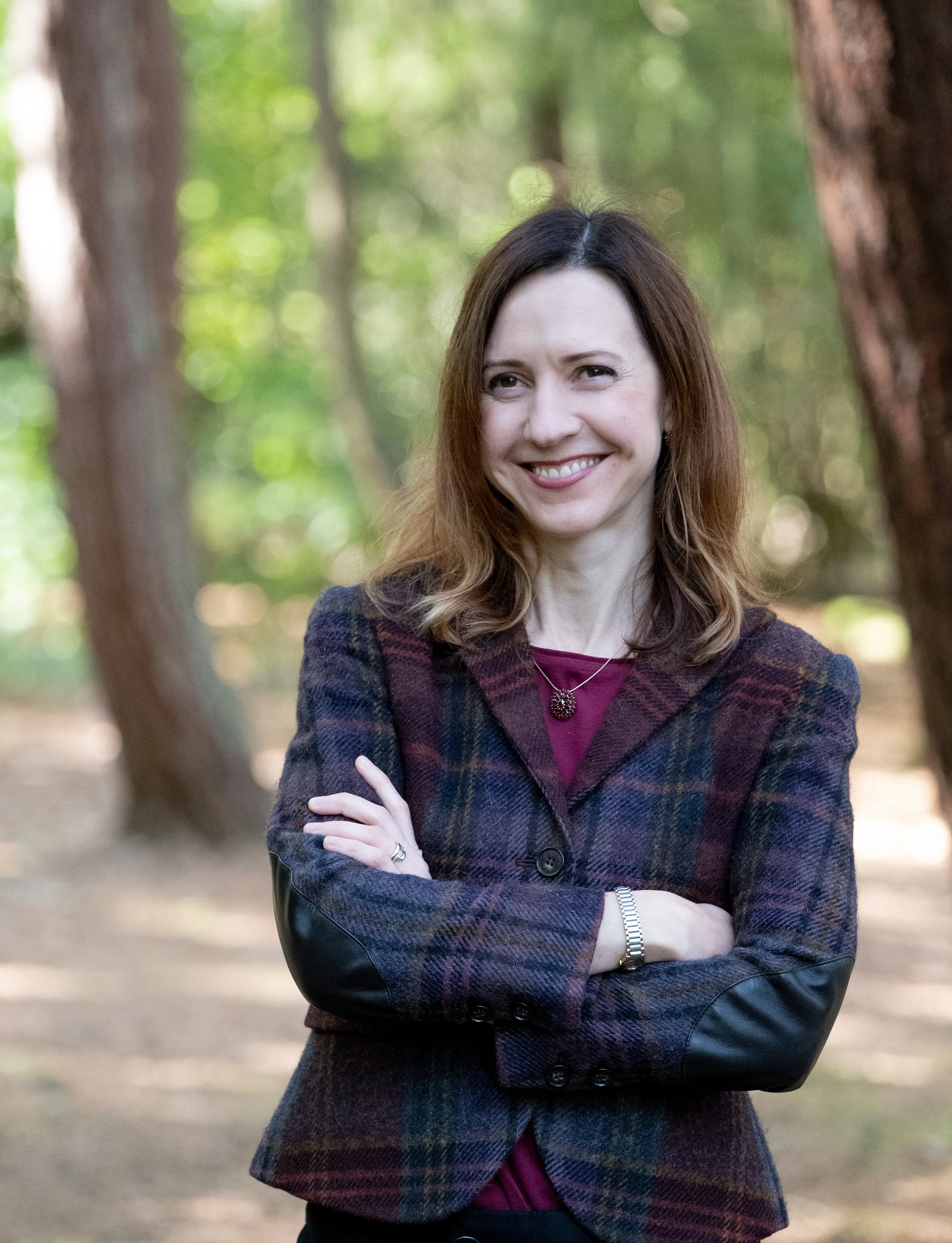 A woman with long brown hair smiling, standing outdoors among trees with blurred green and brown foliage in the background. She is wearing a purple top and a dark plaid blazer with leather sleeves, with arms crossed and wearing jewelry.