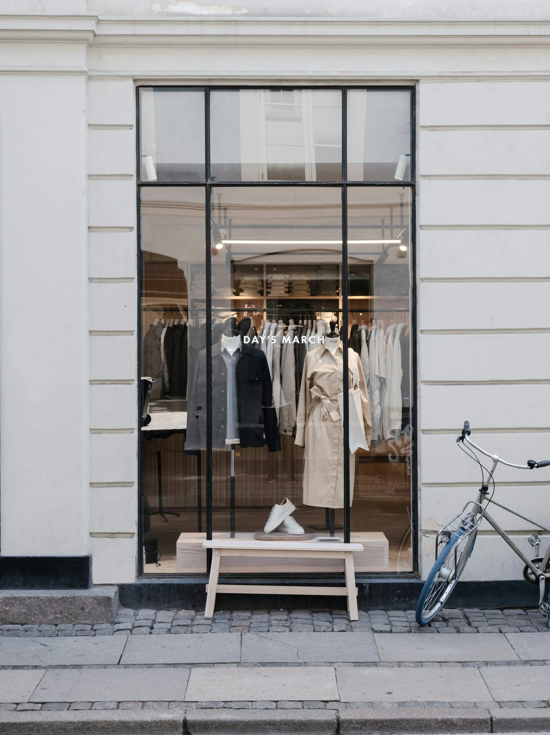 Store front with large window displaying mannequins dressed in neutral and dark clothing, inside shelving with folded fabrics, and the name "DAYS MARCH" on the window. A silver bicycle is parked on the sidewalk to the right.