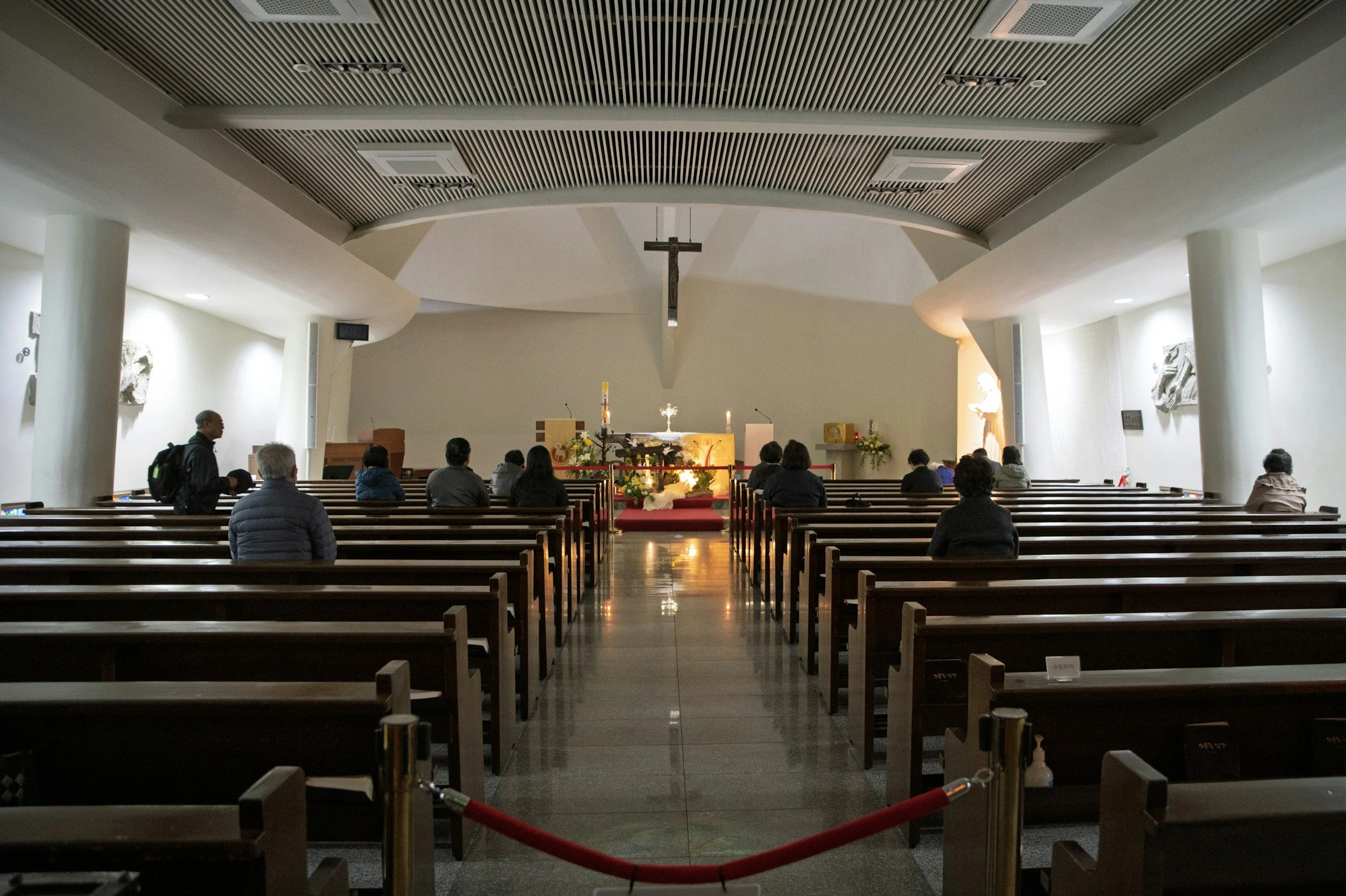 Interior of a church or chapel with pews, an altar with flowers and candles, a crucifix hanging from the ceiling, and a few congregants seated and standing.