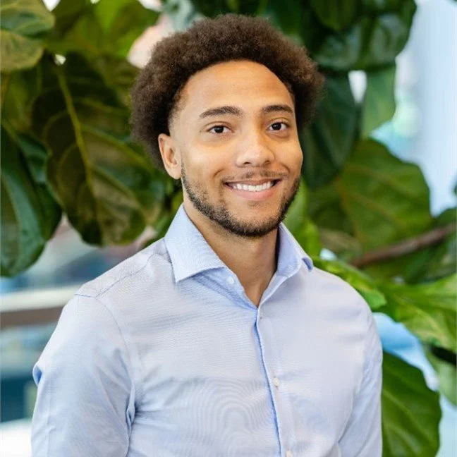 Portrait of a young man with curly hair, wearing a light blue dress shirt, standing in front of green houseplants and smiling.