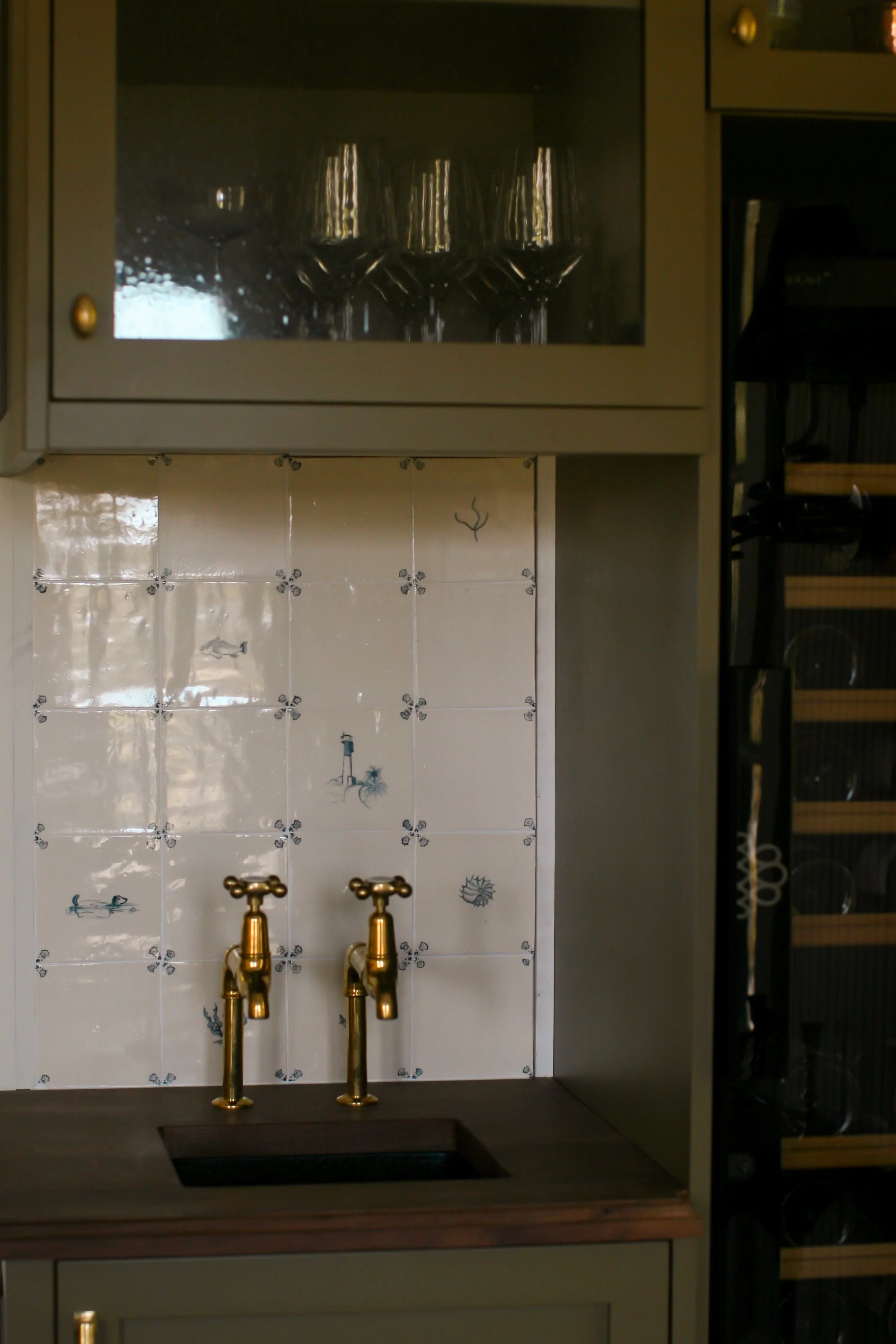 A kitchen sink with two gold faucets in front of a white tile backsplash with blue decorative elements, including fish and a palm tree, and a black countertop. Behind the sink, there is a cabinet with glass doors displaying wine glasses inside.