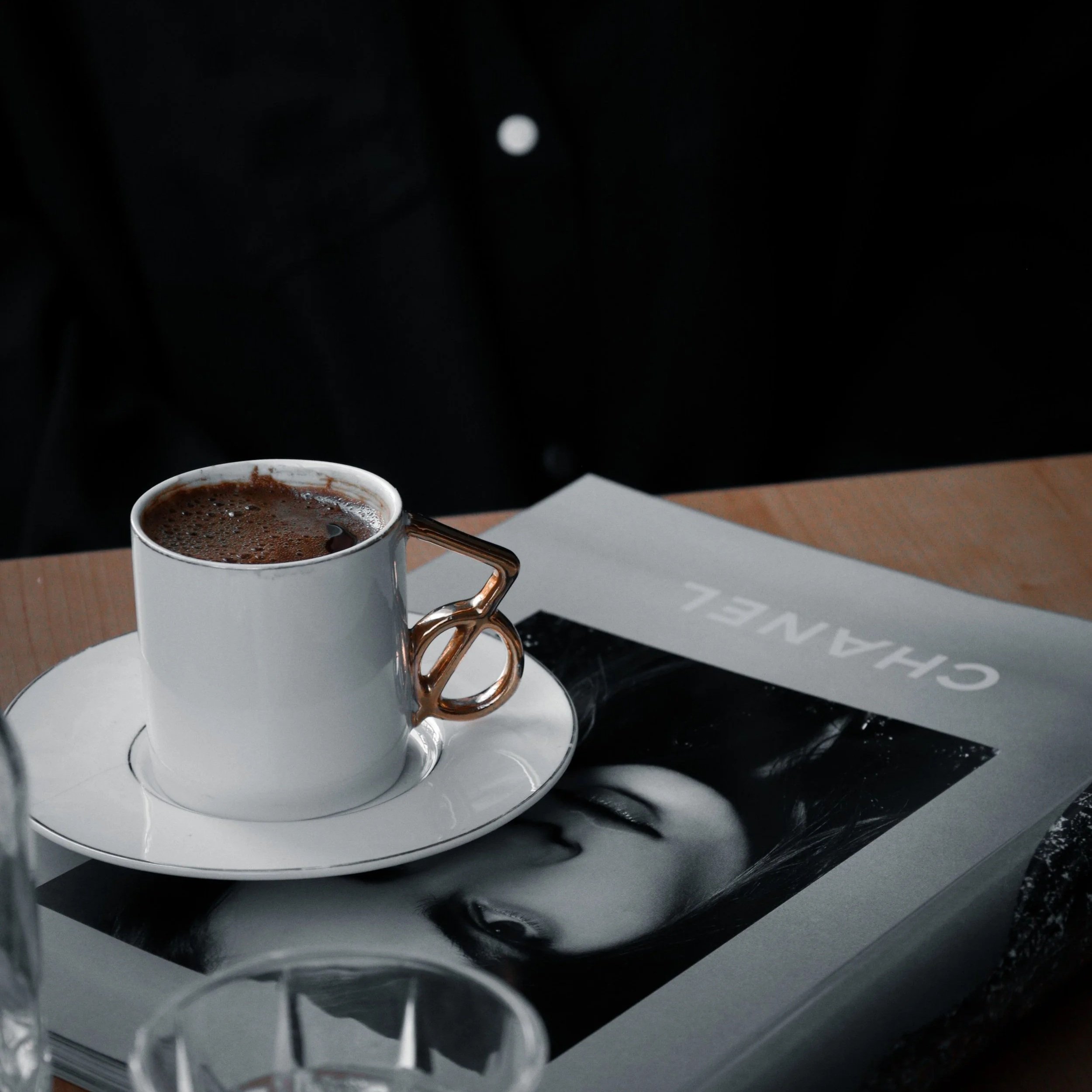 Close-up of a white coffee mug filled with hot chocolate, placed on a white saucer on a table, with black background and a menu or paper nearby.
