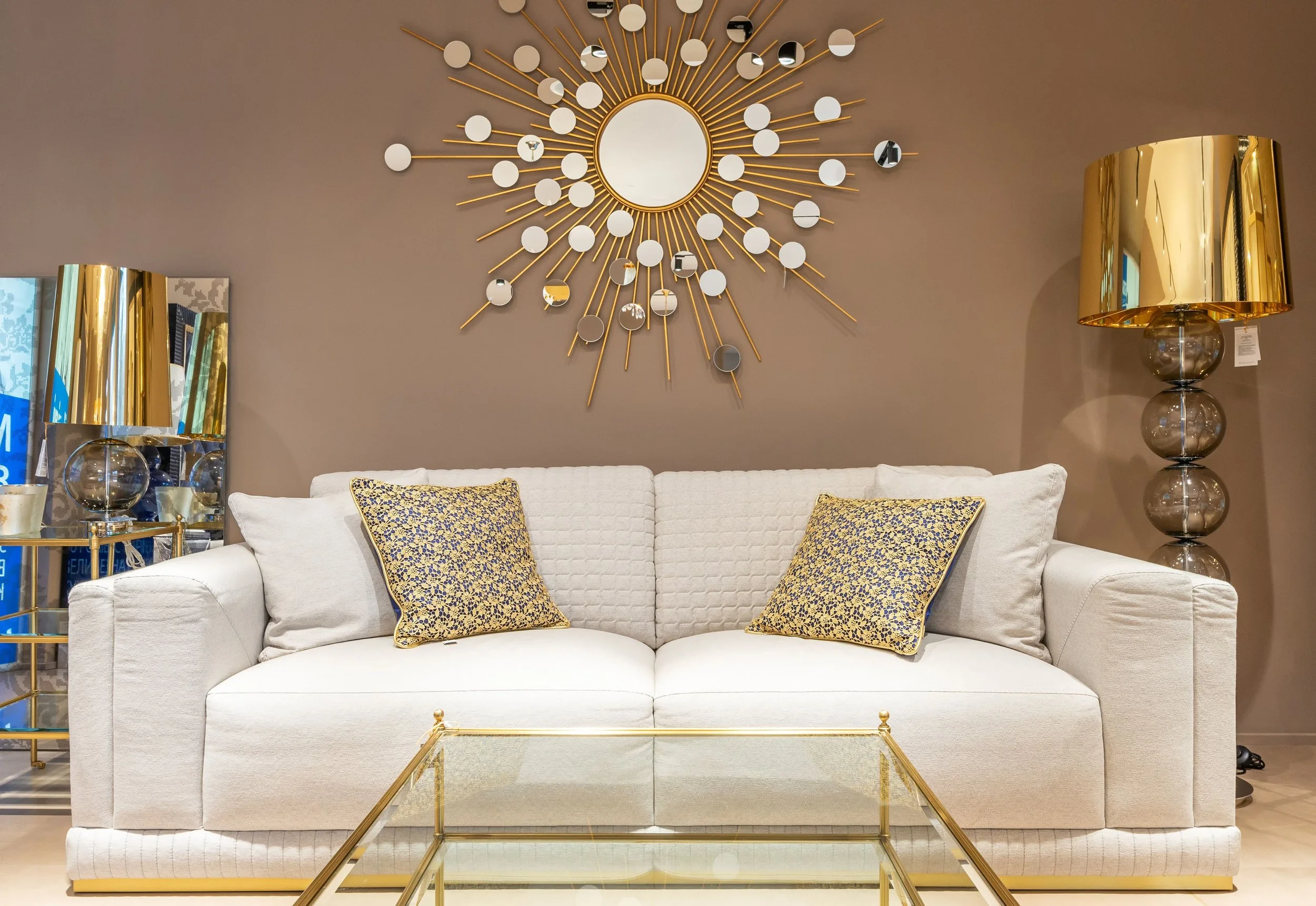 Living room with a white sofa, decorative pillows, a glass coffee table, a gold and glass table lamp, and a sunburst mirror on a beige wall.