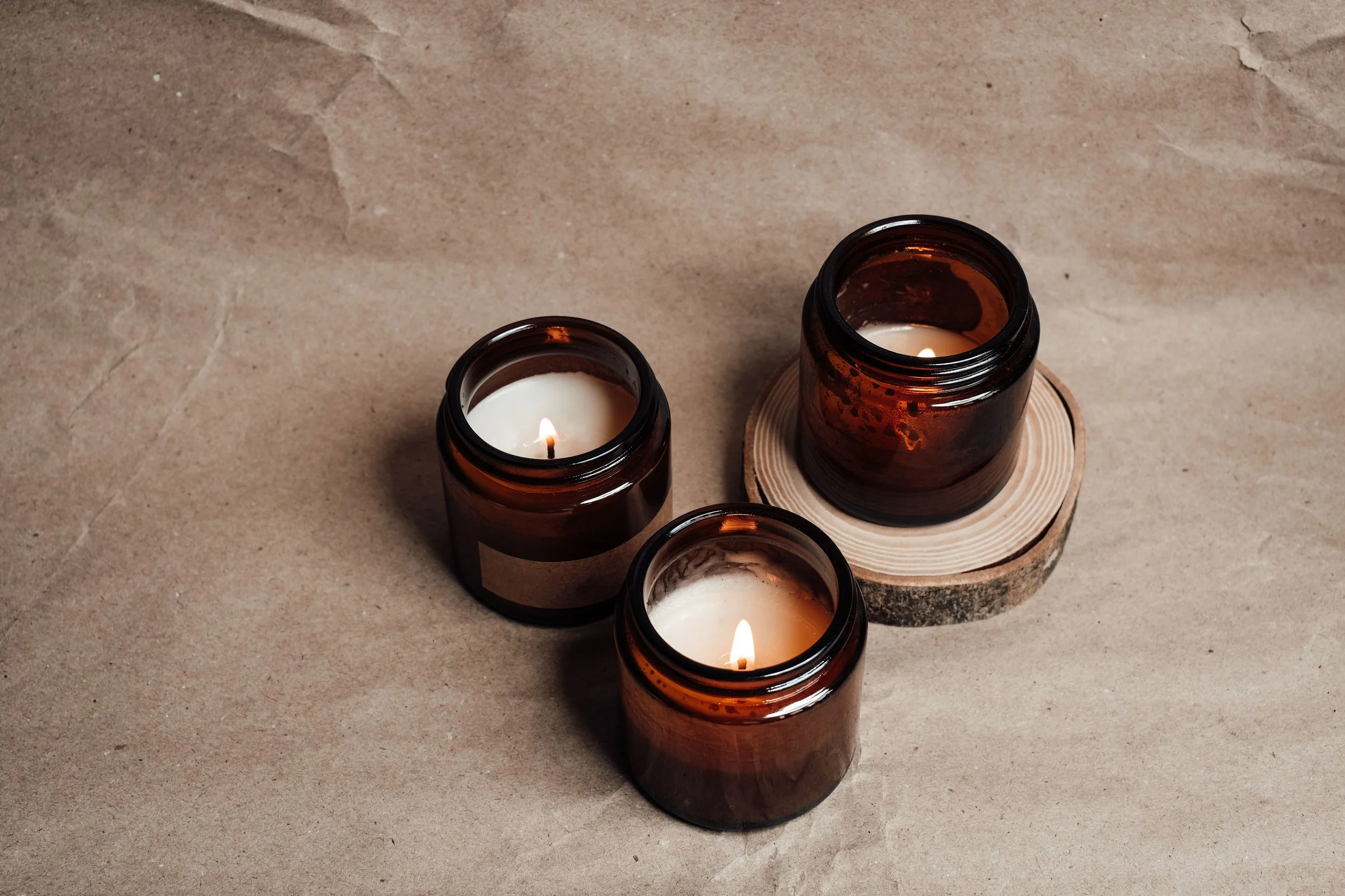 Four lit candles in brown glass jars placed on a wooden coaster on a beige surface.