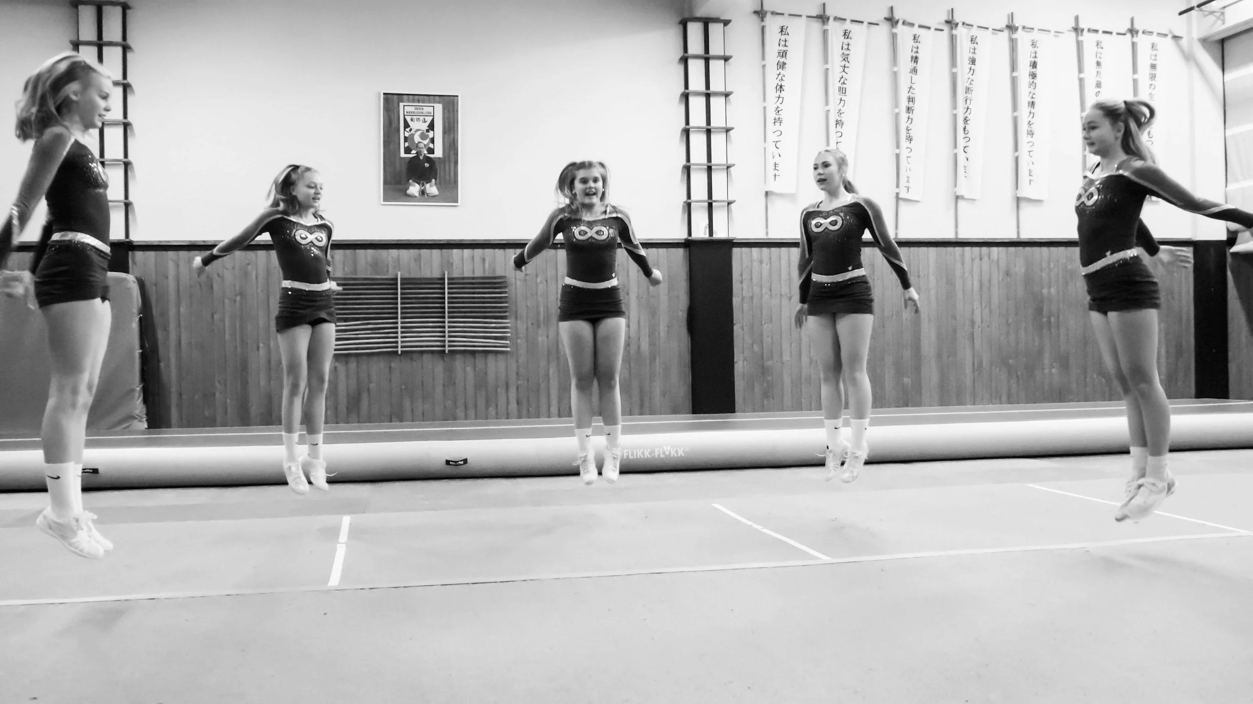 Five young girls in gymnastics leotards jumping simultaneously during a practice session in a gymnasium.