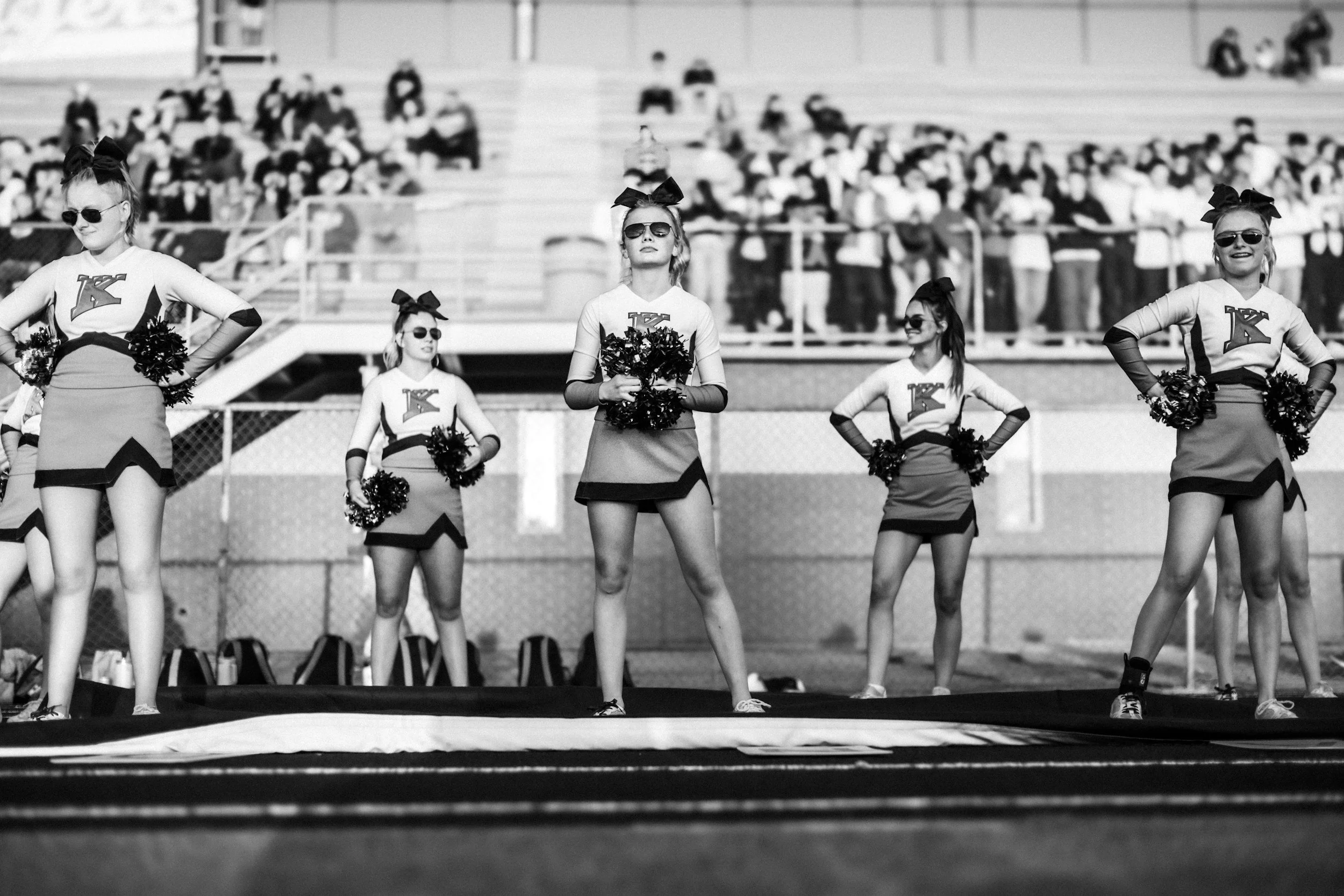 Cheerleading. A black-and-white photo of cheerleaders standing on a football field with their hands on their hips, holding pom-poms. They are wearing matching uniforms and large bows in their hair, with a crowd in the background.
