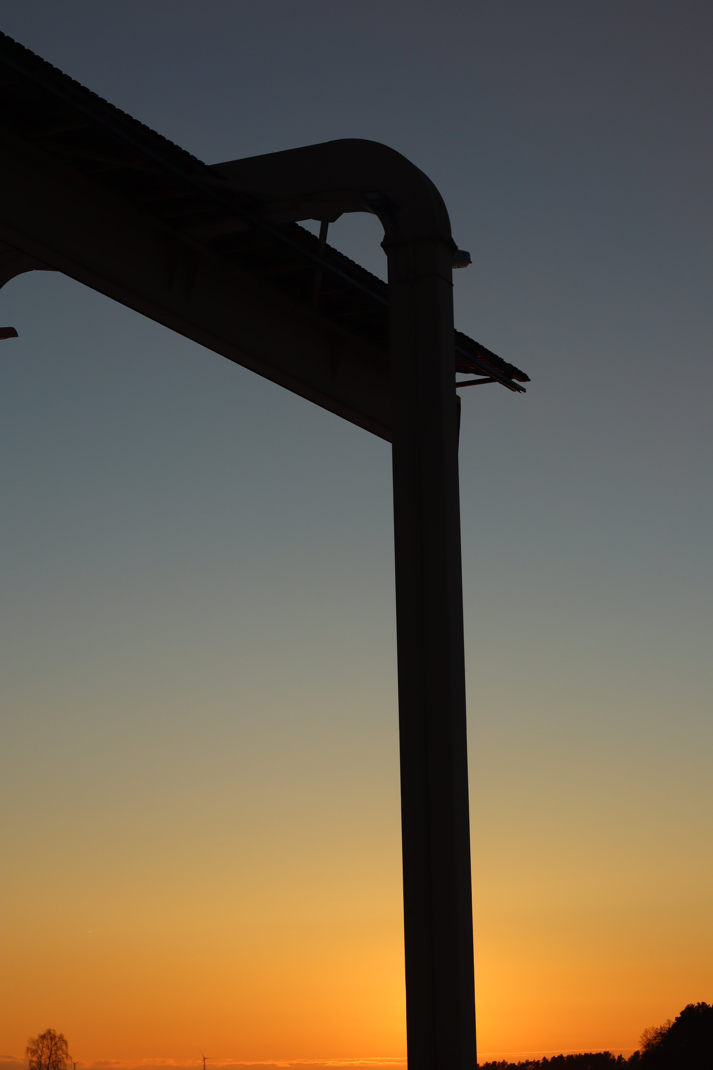 Silhouette of a tall wind turbine against a sunset sky with a gradient from yellow to dark blue, with trees in the distance.