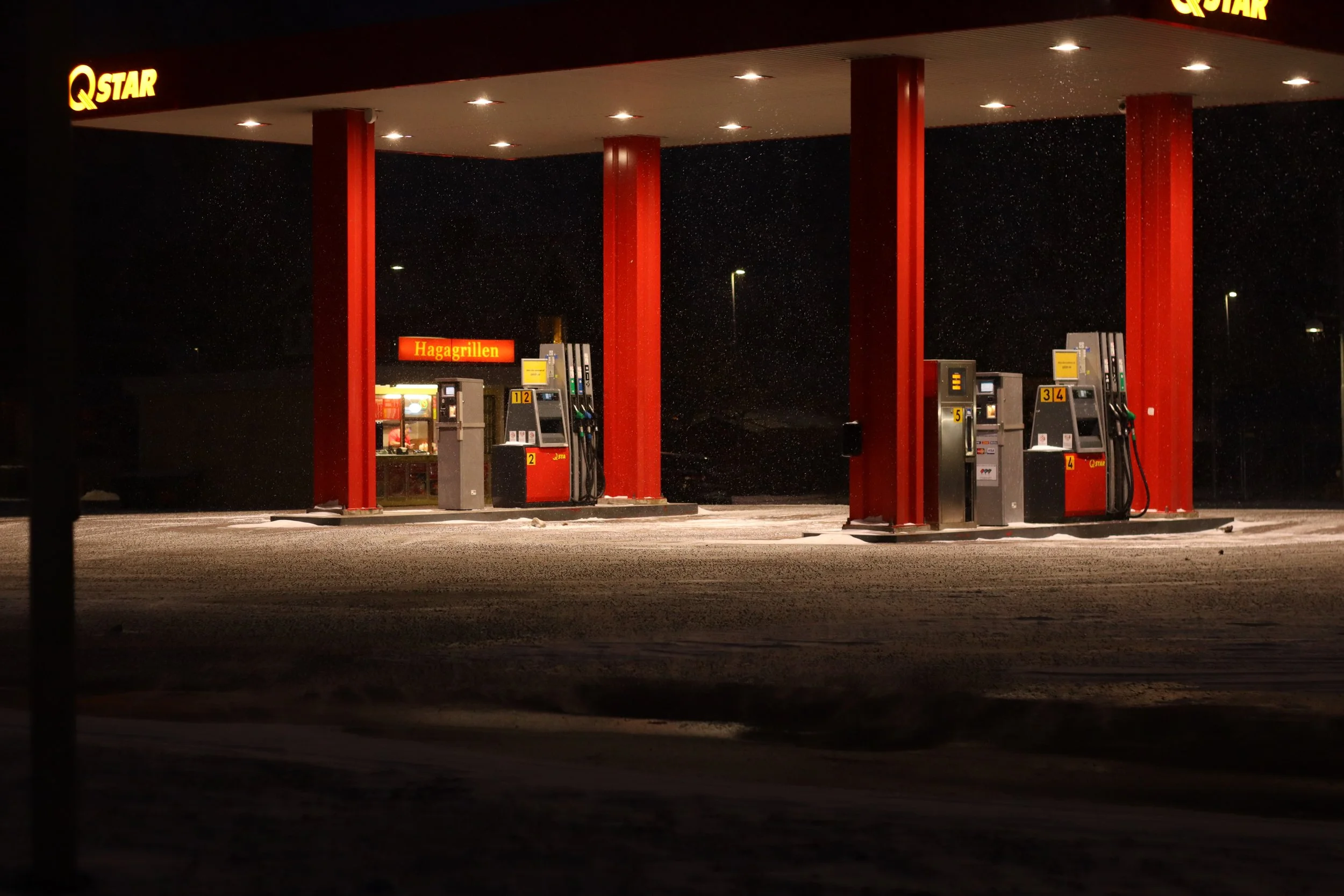 An image of a gas station at night with pumps under a roof, illuminated by ceiling lights, and a sign that reads 'Hagagrillen' in the background. Snow is falling lightly on the ground.