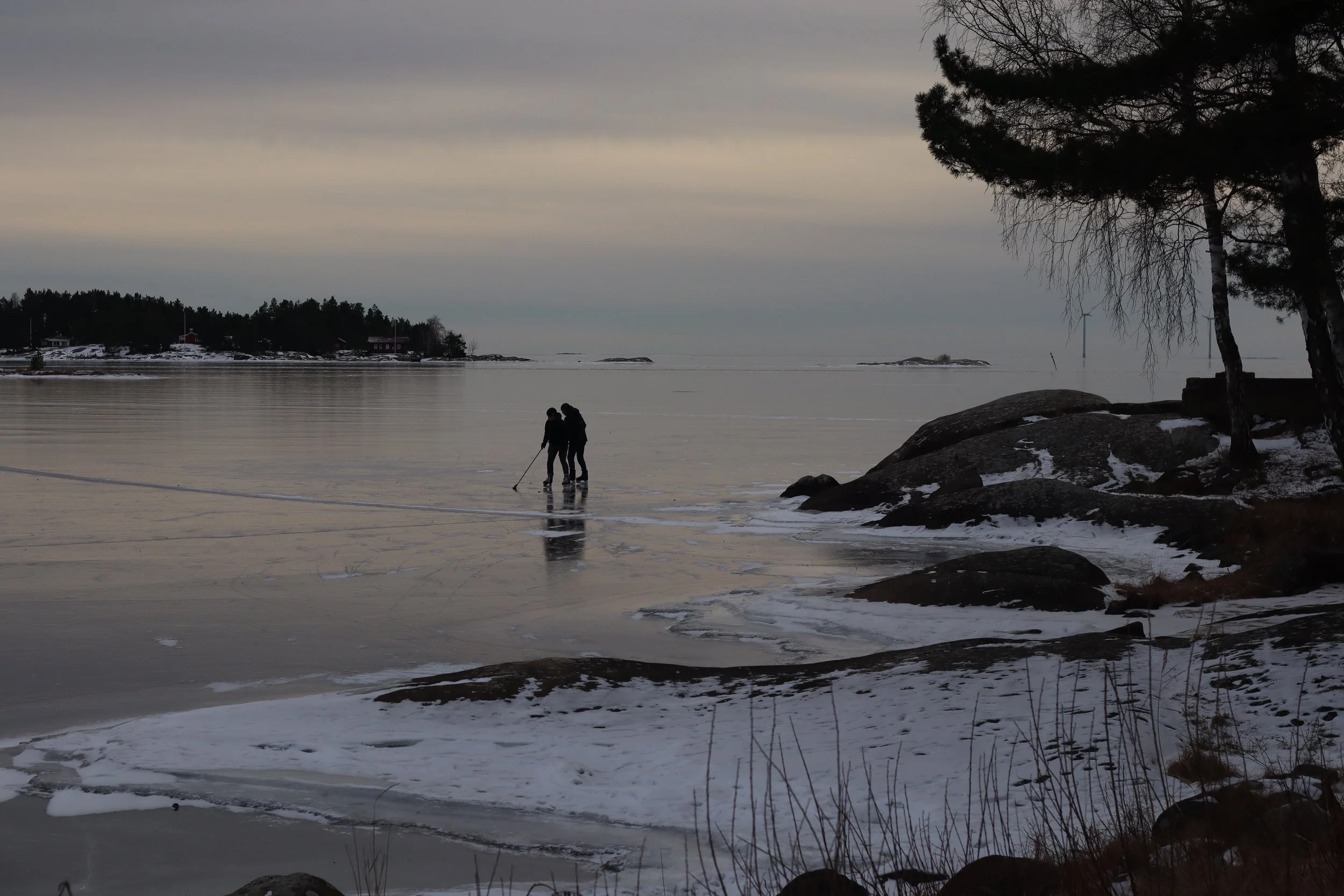 Two people walking on a frozen lake near the shoreline with rocks and trees, overcast sky, distant houses, and wind turbines in the background.