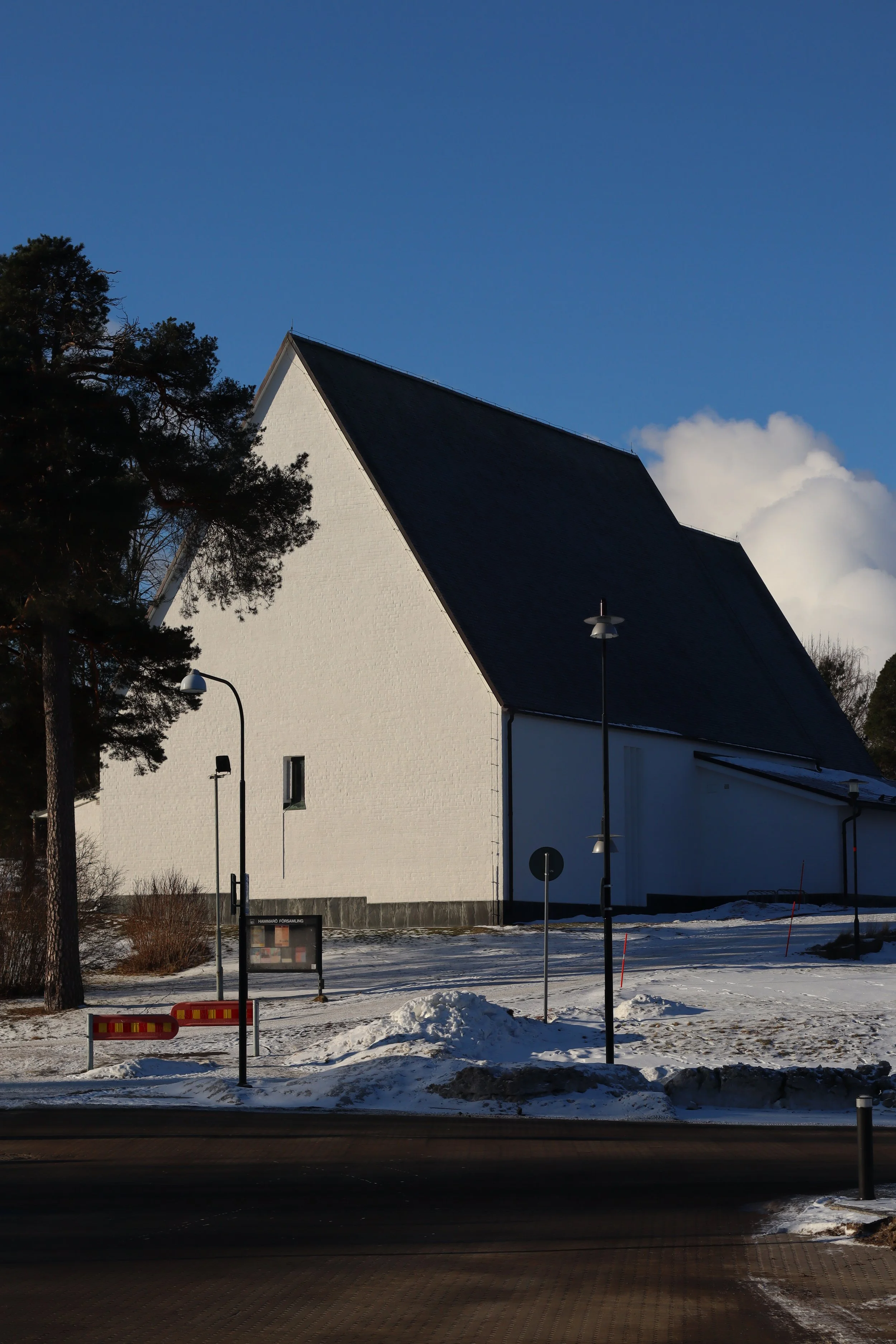 A white church with a black roof in a snowy landscape, trees on one side, and street signs and lamps in the foreground, winter season with shadows on the snow.