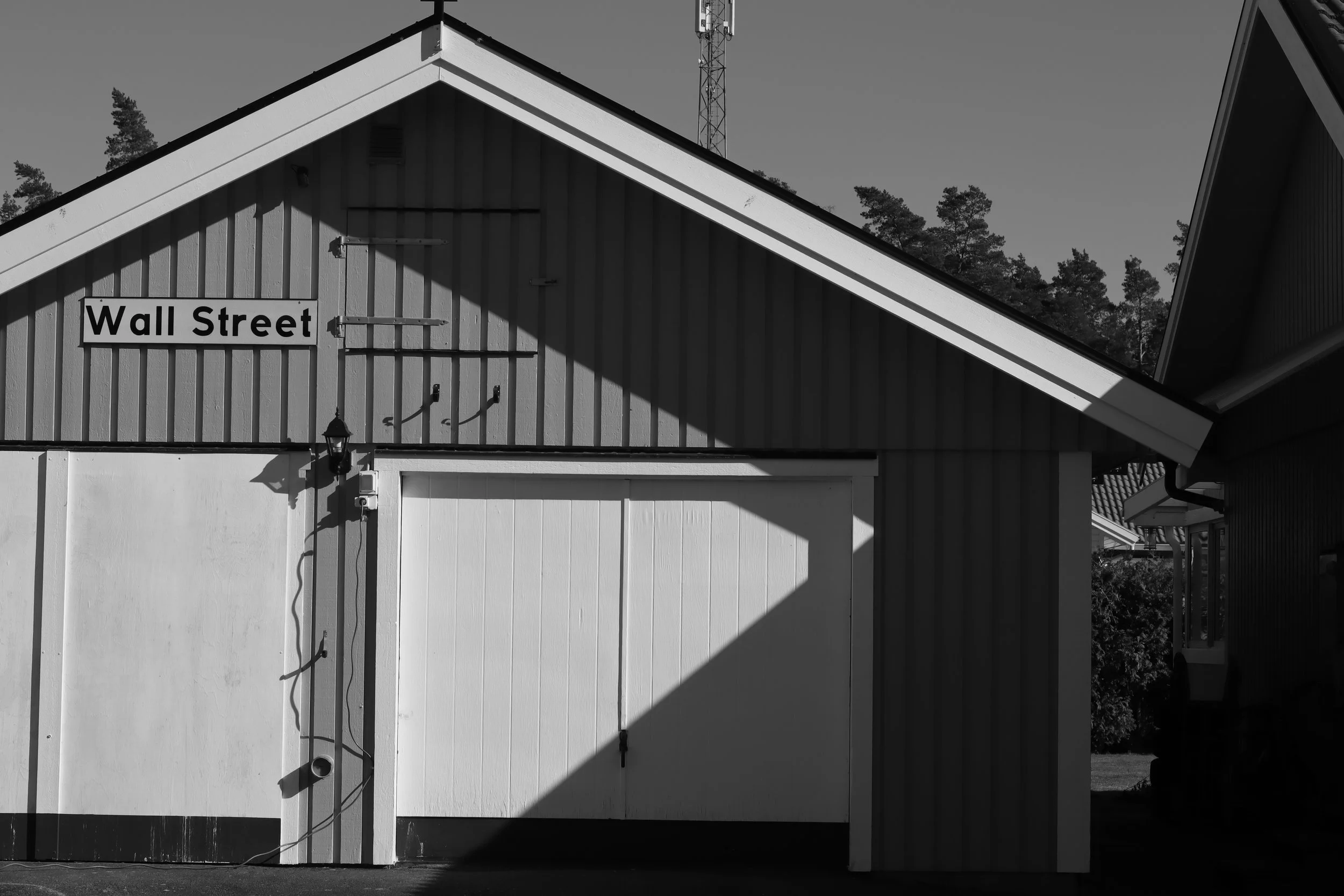 Front of a building with a sign that reads "Wall Street". The building has a sloped roof and exterior walls with vertical siding. There are shadows cast on the building, and in the background, there are trees and a tall communication tower.