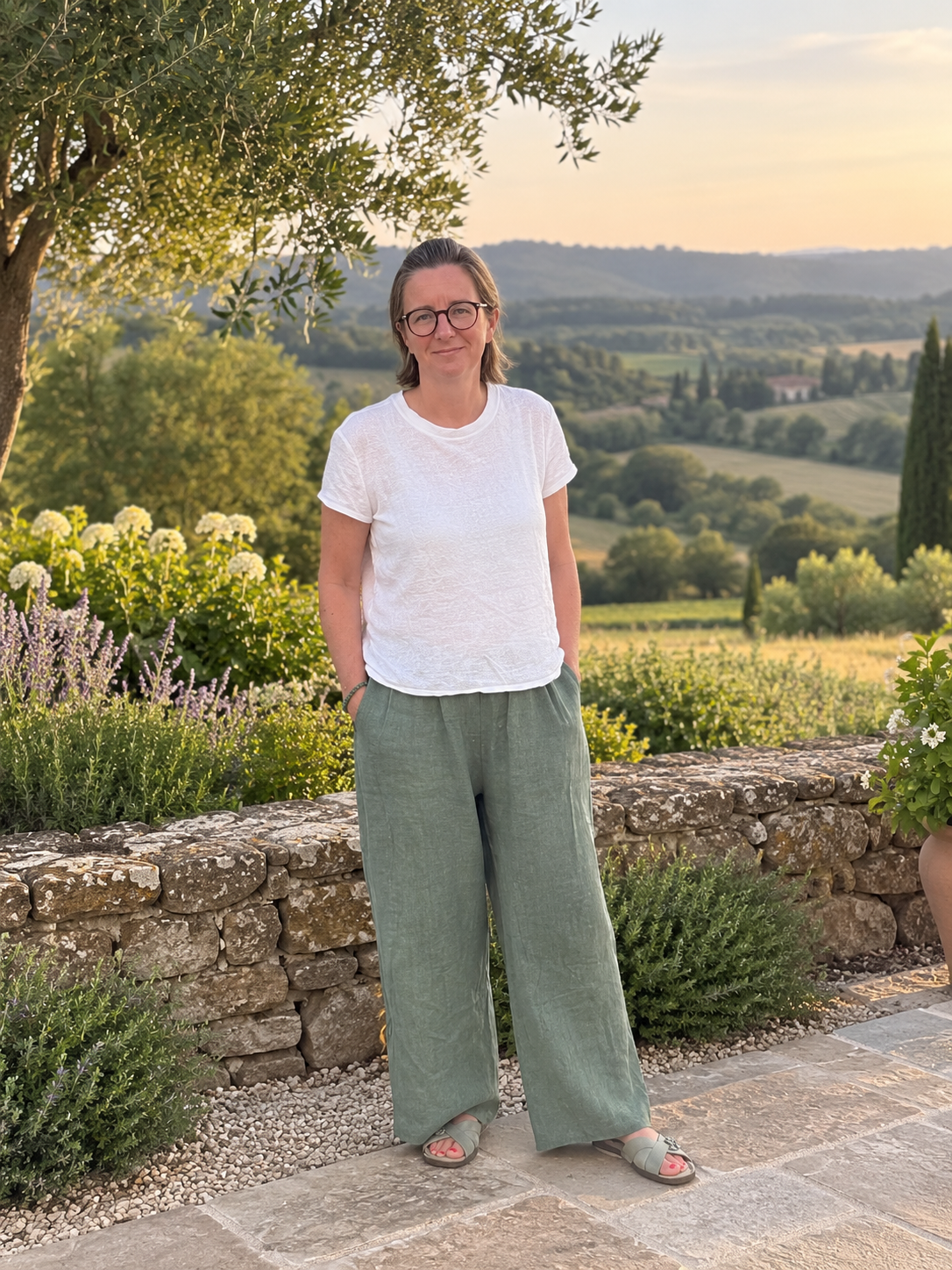A woman with glasses wearing a white t-shirt, green wide-leg pants, and sandals standing outdoors in a garden with a stone wall and lush landscape of rolling hills and trees in the background during sunset.