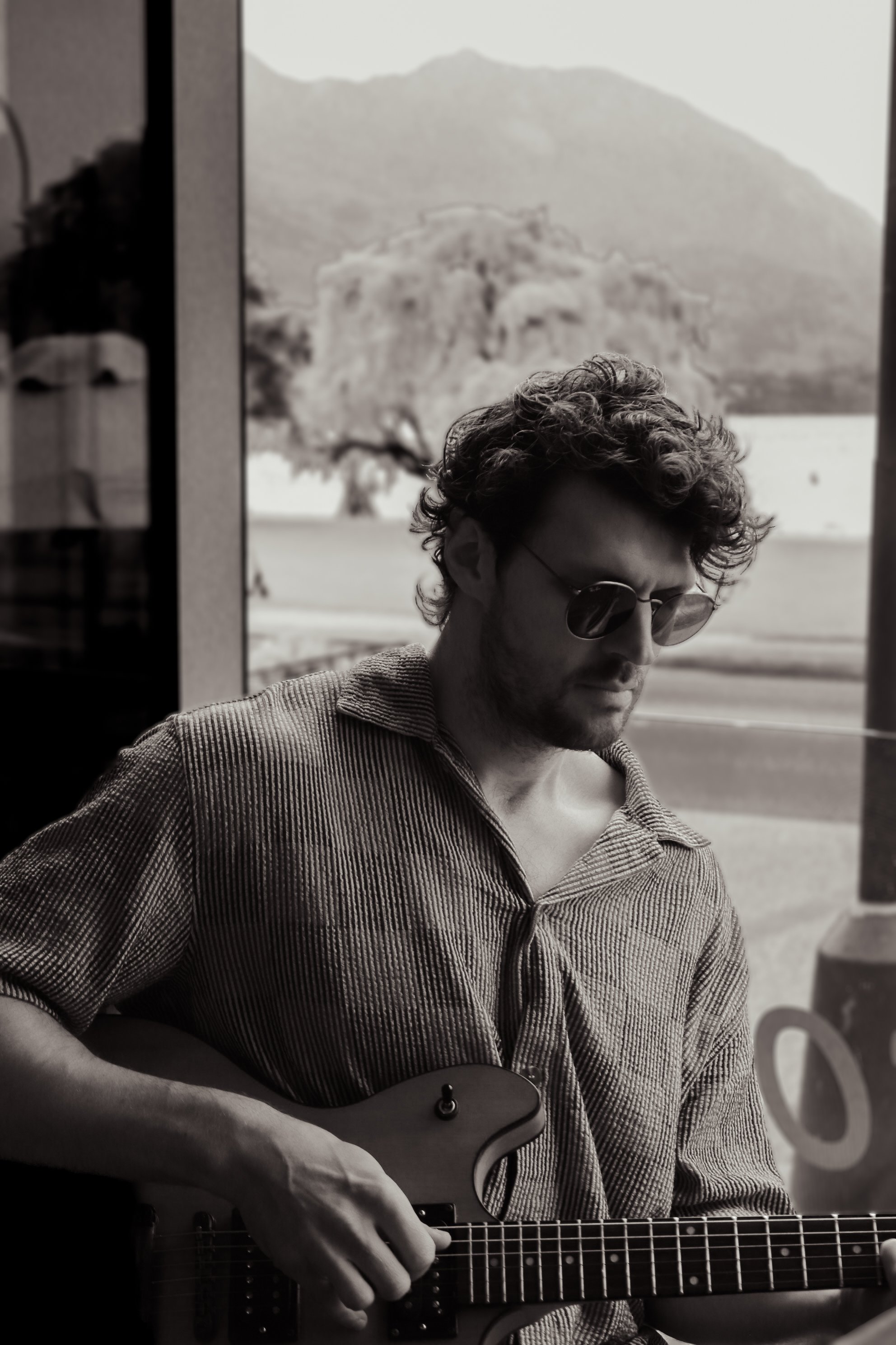 A man with curly hair and sunglasses playing an electric guitar indoors near a large window with a mountain and tree landscape in the background.