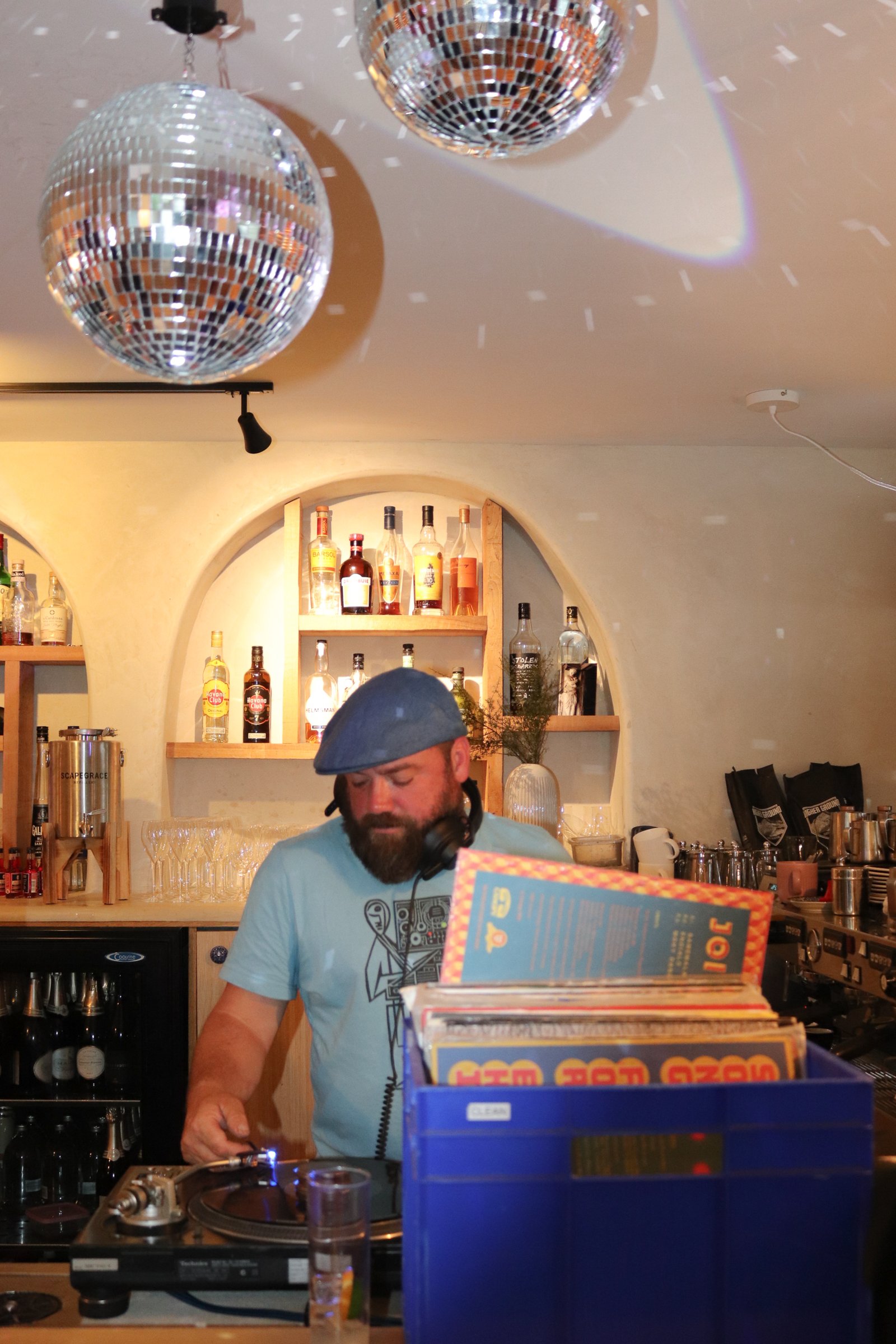 A DJ with a beard wearing a blue beret and headphones around his neck, standing behind a record player, in a bar with shelves of liquor bottles in the background, illuminated by disco ball reflections on the ceiling.