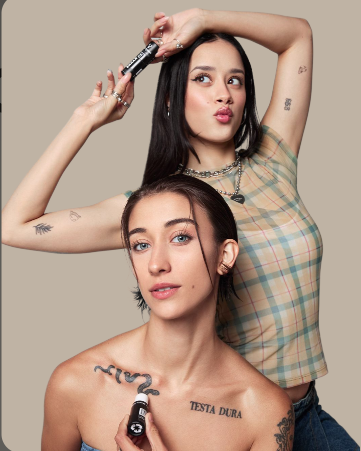Two women with tattoos and makeup, one standing behind the other, holding makeup products and looking at the camera on a neutral background.