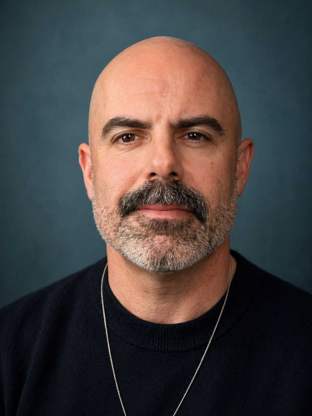A close-up portrait of a middle-aged man with a bald head, brown eyes, and a salt-and-pepper beard and mustache. He is wearing a black shirt and a silver chain necklace, with a plain dark background.