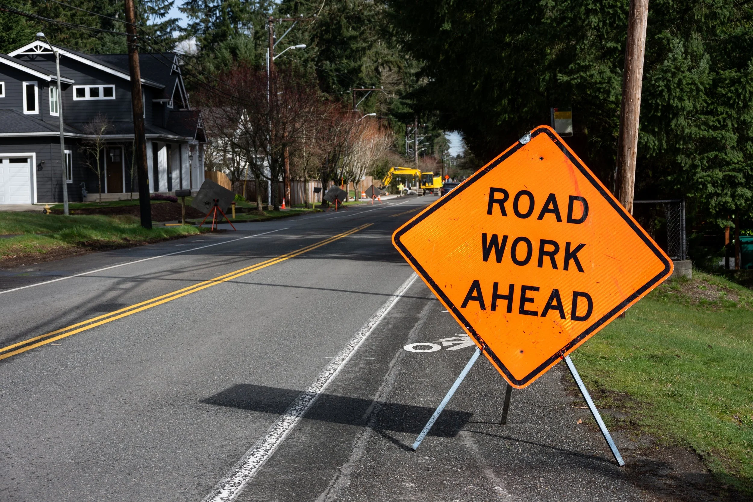 Orange road work ahead sign on a folding stand blocking part of a residential street, with construction equipment and cones further down the road, houses and trees lining the street.