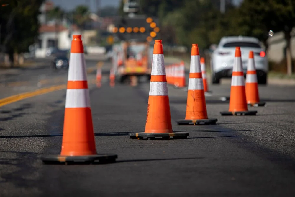 Orange traffic cones block off a section of a road under construction, with a construction vehicle and a white SUV in the background.