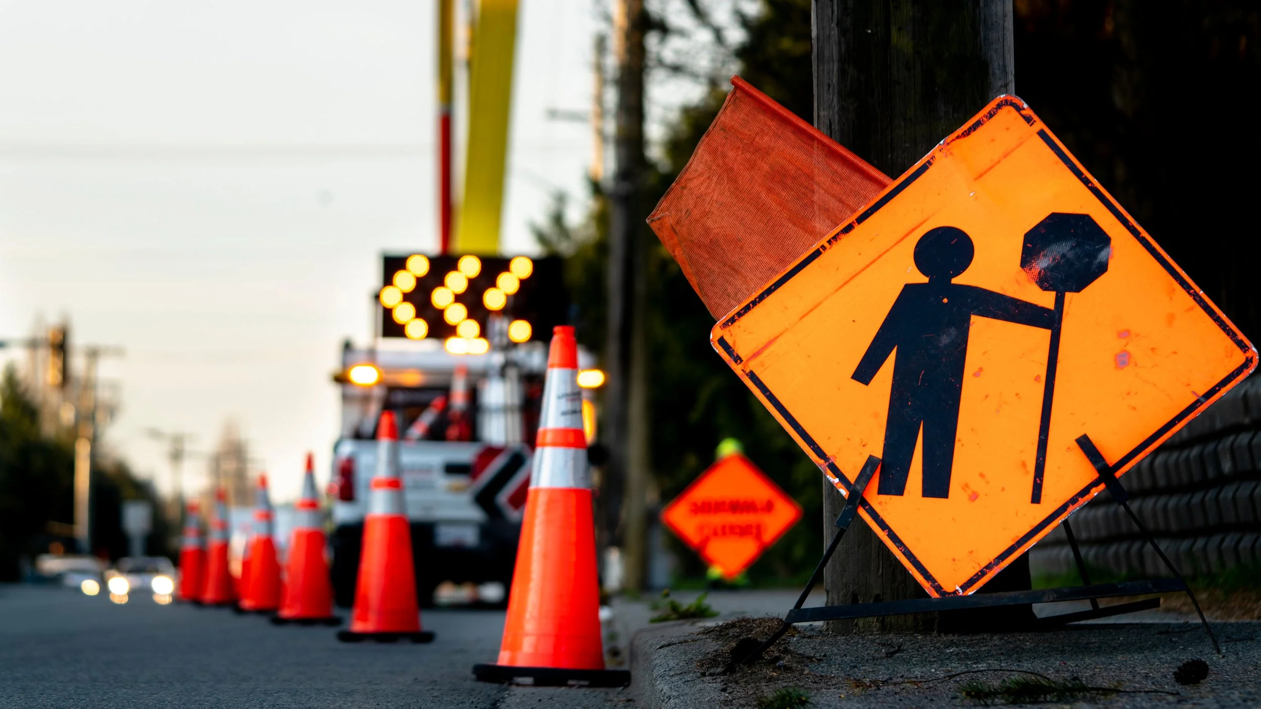 Road construction zone with orange traffic cones, warning signs, and a worker truck with flashing lights in the background.