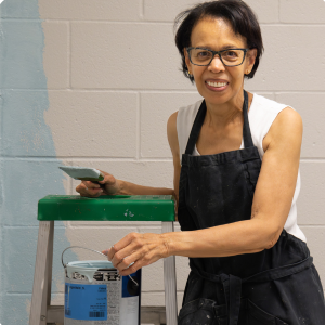 Smiling woman with glasses, wearing a white shirt and black apron, holding a paintbrush and a paint roller, standing next to a paint can on a step ladder.