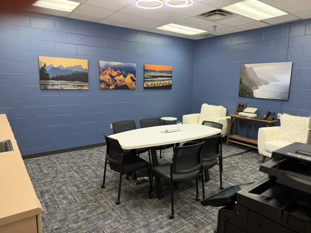 A conference room with a blue wall decorated with four landscape paintings, a white oval table surrounded by black chairs, and white armchairs with a small side table on the right.