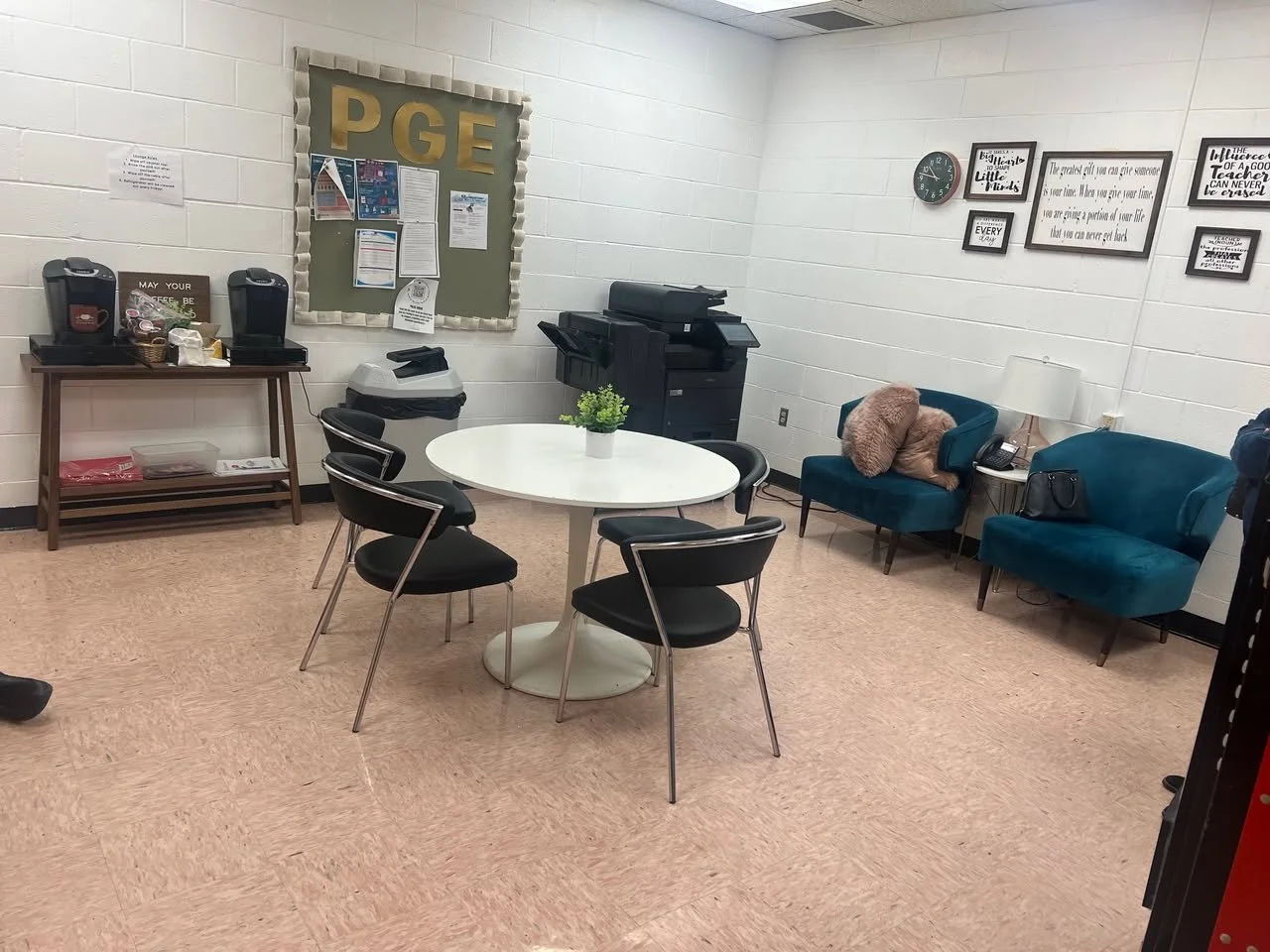 A small waiting area with a round white table, four black chairs, two teal armchairs, a beige pillow on one, a white lamp on a side table, a black handbag, an indoor plant, a bulletin board on white brick wall, framed quotes, clock, a copier, and two coffee machines.