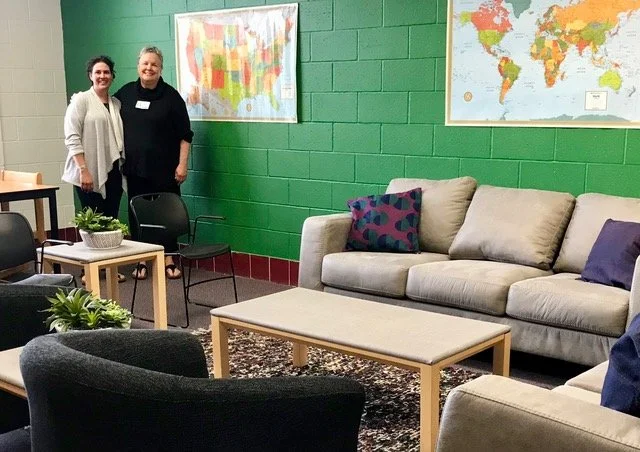 Two women standing in a colorful office with green brick walls, world maps on the wall, and a beige couch with purple and patterned pillows, surrounded by black and beige chairs, small tables, and potted plants.
