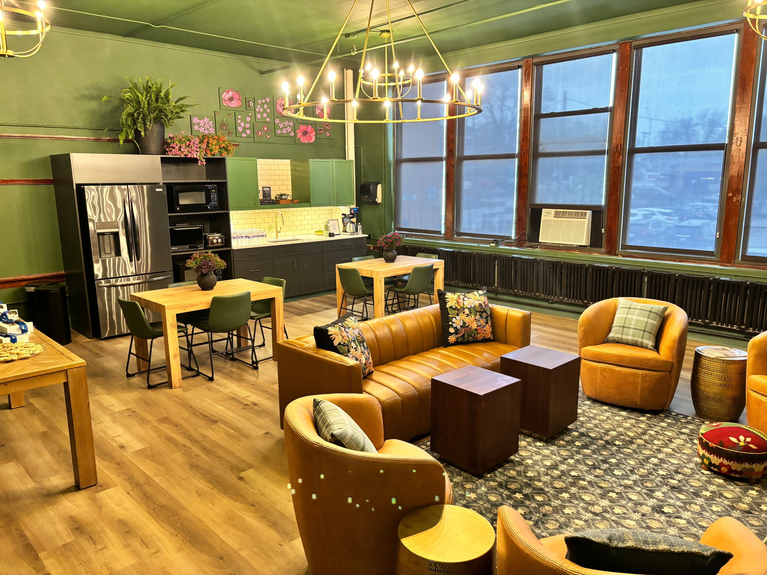 A cozy, modern kitchen and living area with green walls and large windows, featuring wooden tables, a leather sofa with floral cushions, a black bookshelf with kitchen appliances, and plants, illuminated by a chandelier.