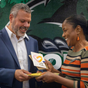 A man in a suit and a woman with short hair in a striped shirt exchanging an award or certificate in front of a colorful mural.
