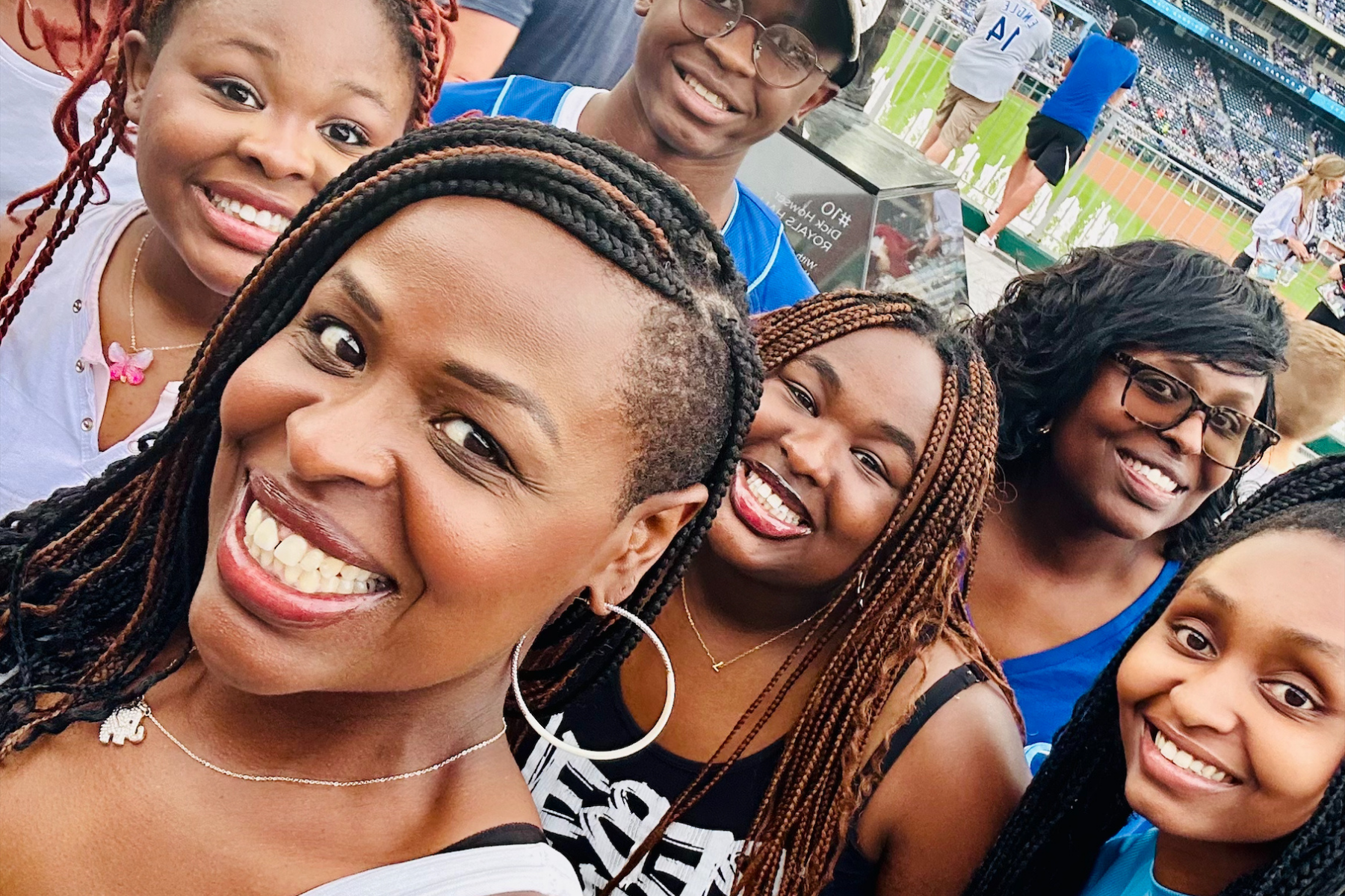 Group of diverse women smiling at a sports stadium, with some wearing glasses, sporting braided hairstyles, and wearing casual attire.