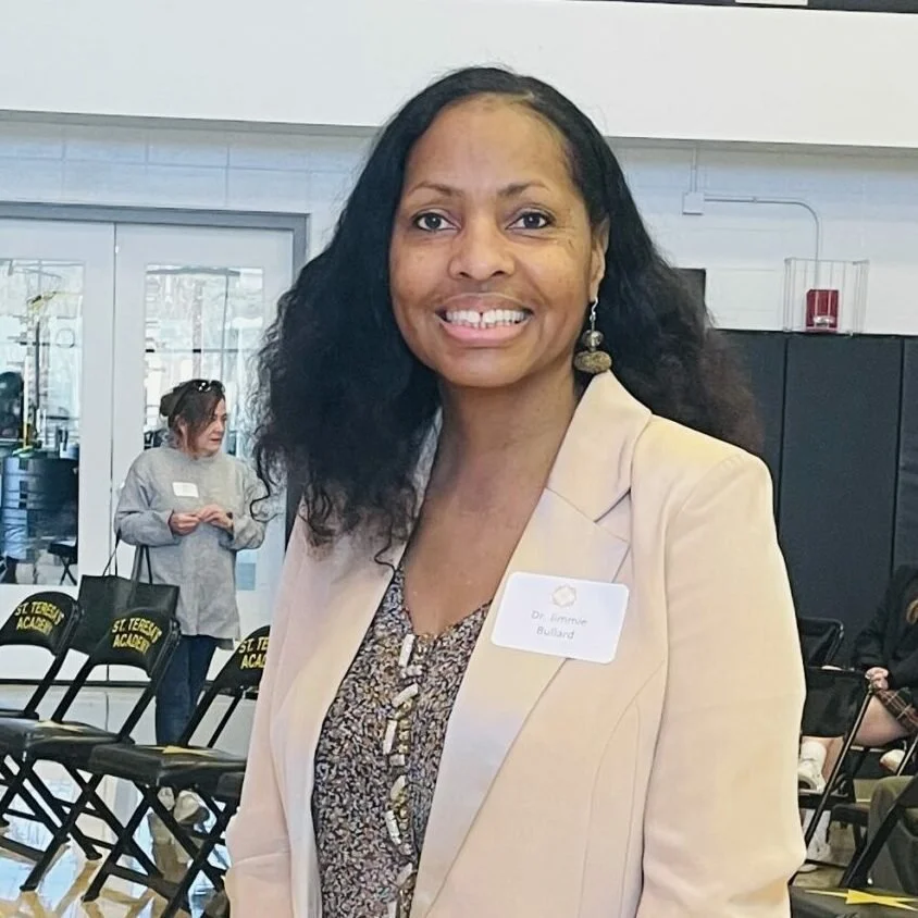 A woman smiling at an indoor event, wearing a beige blazer and a patterned top, with a name tag that reads 'Dr. Symme Buillard'. There are chairs and people in the background.